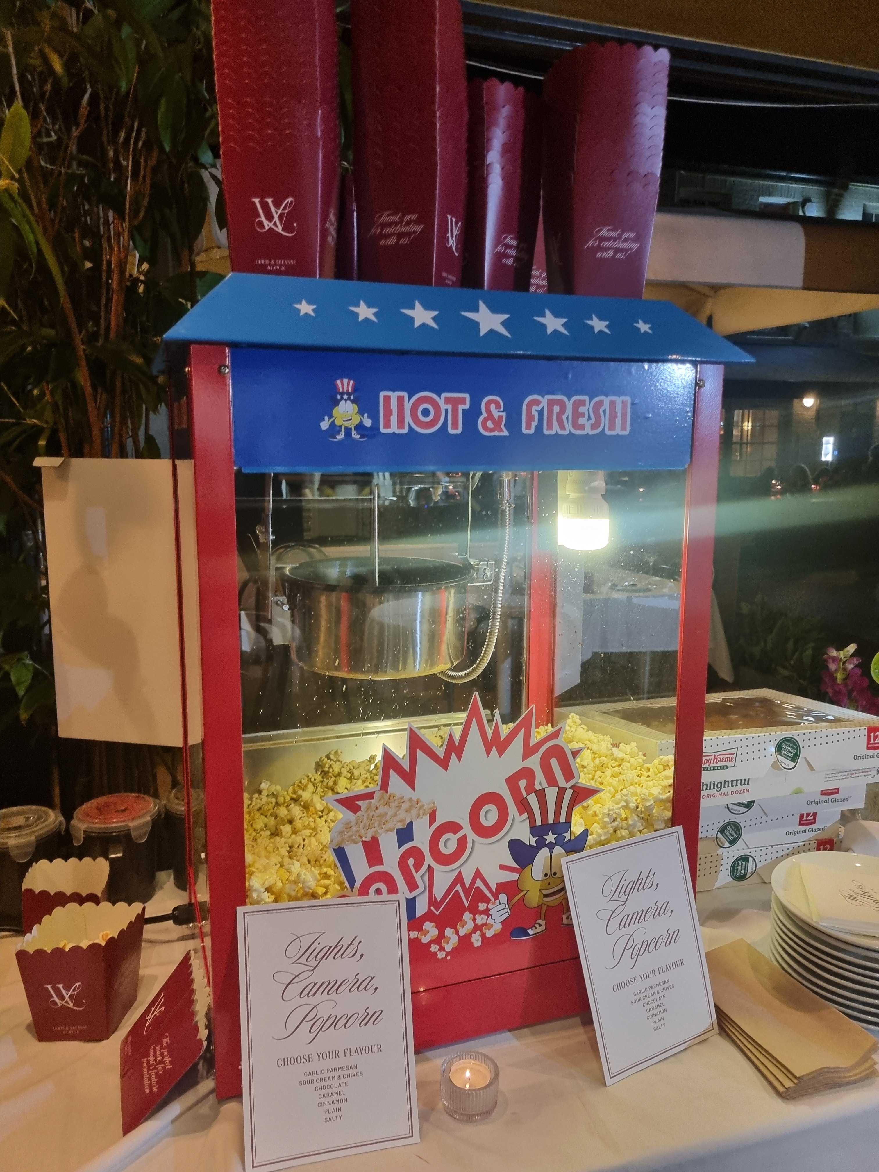 Bright night catering display with a blue-red popcorn machine, warm light, flavor cards, stacked boxes, and polished event buffet service.