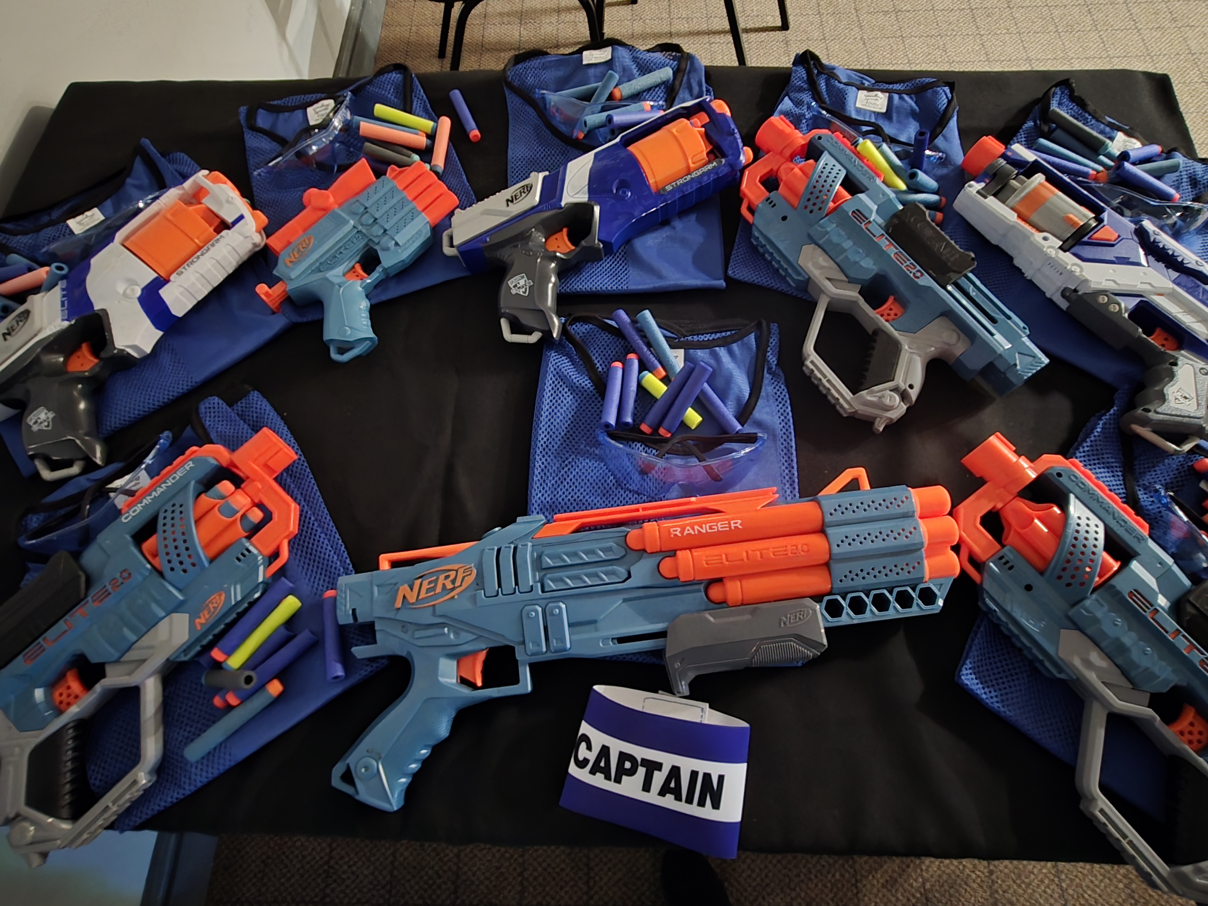 A festive setup features vibrant Nerf blasters in blue and orange, organized around colorful ammo-filled mesh pouches on a black table.