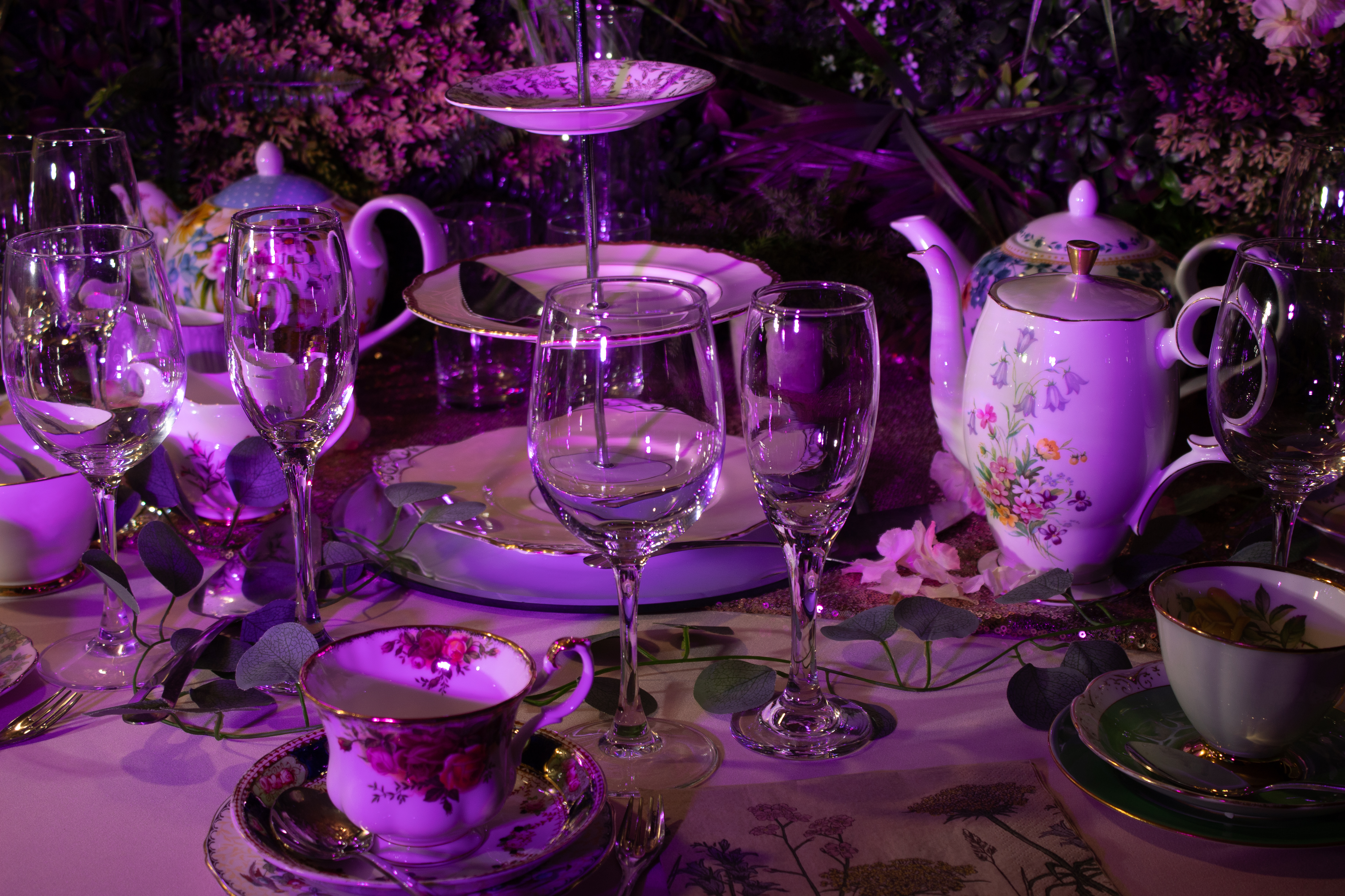 An elegant table setting features floral-patterned china, crystal glasses, and a tiered stand, all under soft purple lighting, perfect for catering events.