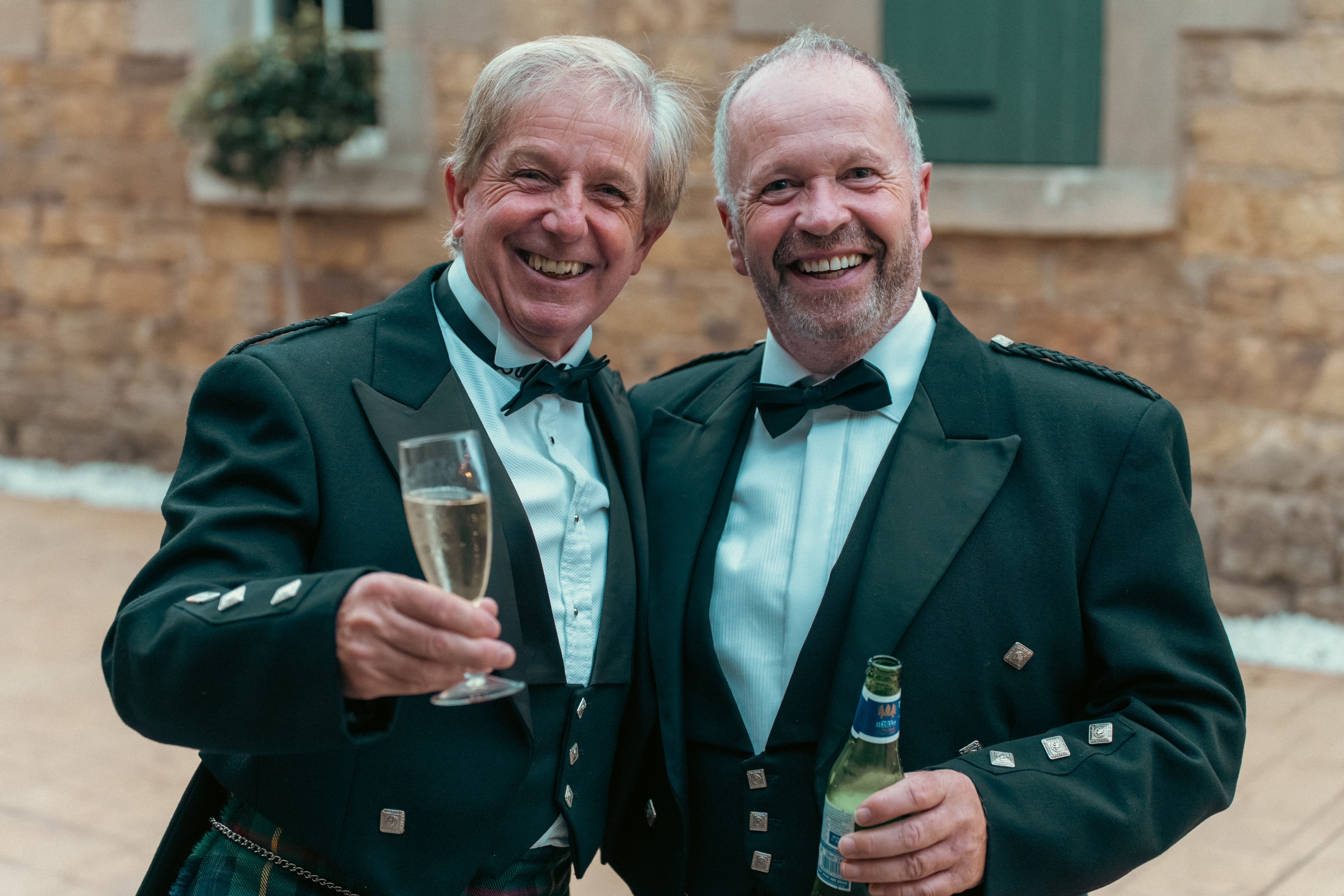 Two jovial gentlemen in formal kilts and tuxedos toast with champagne and beer, exuding warmth in a rustic stone backdrop.