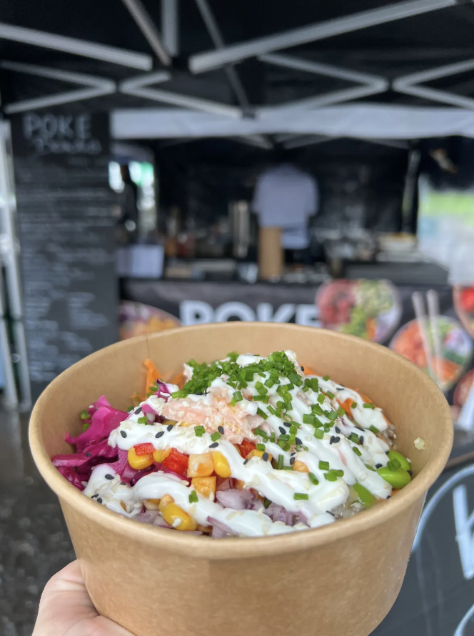 Colorful poke bowl in a kraft paper cup, topped with salmon, avocado, corn, pickled cabbage, and creamy mayo, at a sleek catering stall.