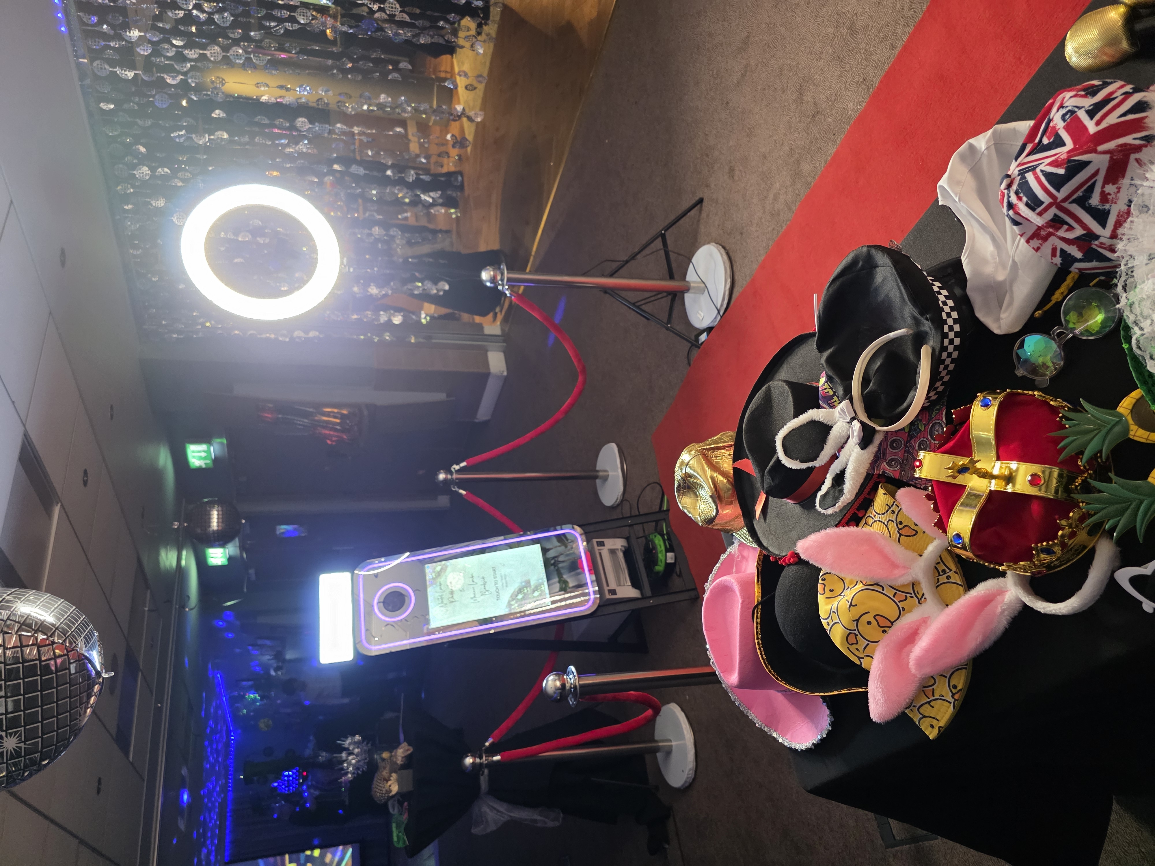 A vibrant event scene features a variety of colorful hats and props on a black table, illuminated by warm lights and disco balls.