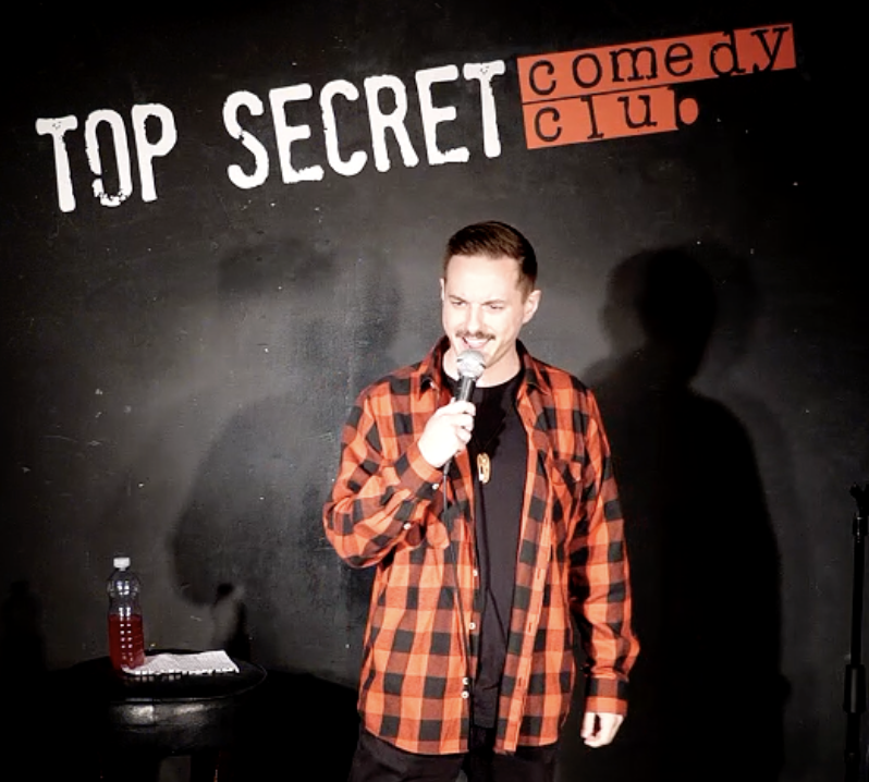 Comedian in orange plaid shirt performs with microphone on a small black stage, Top Secret Comedy Club sign above, moody spotlight.