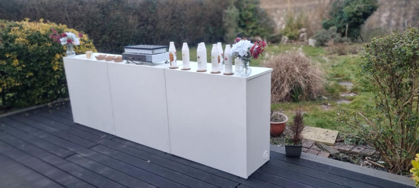 A sleek white catering table showcases glass vases with flowers, bottles capped on coasters, and a cooking station, set in soft daylight.