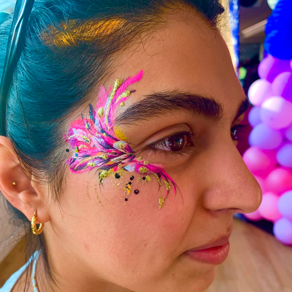 Close-up of a woman with neon pink and gold face paint, gold hoop earrings, and purple balloon decor, under bright event lighting.
