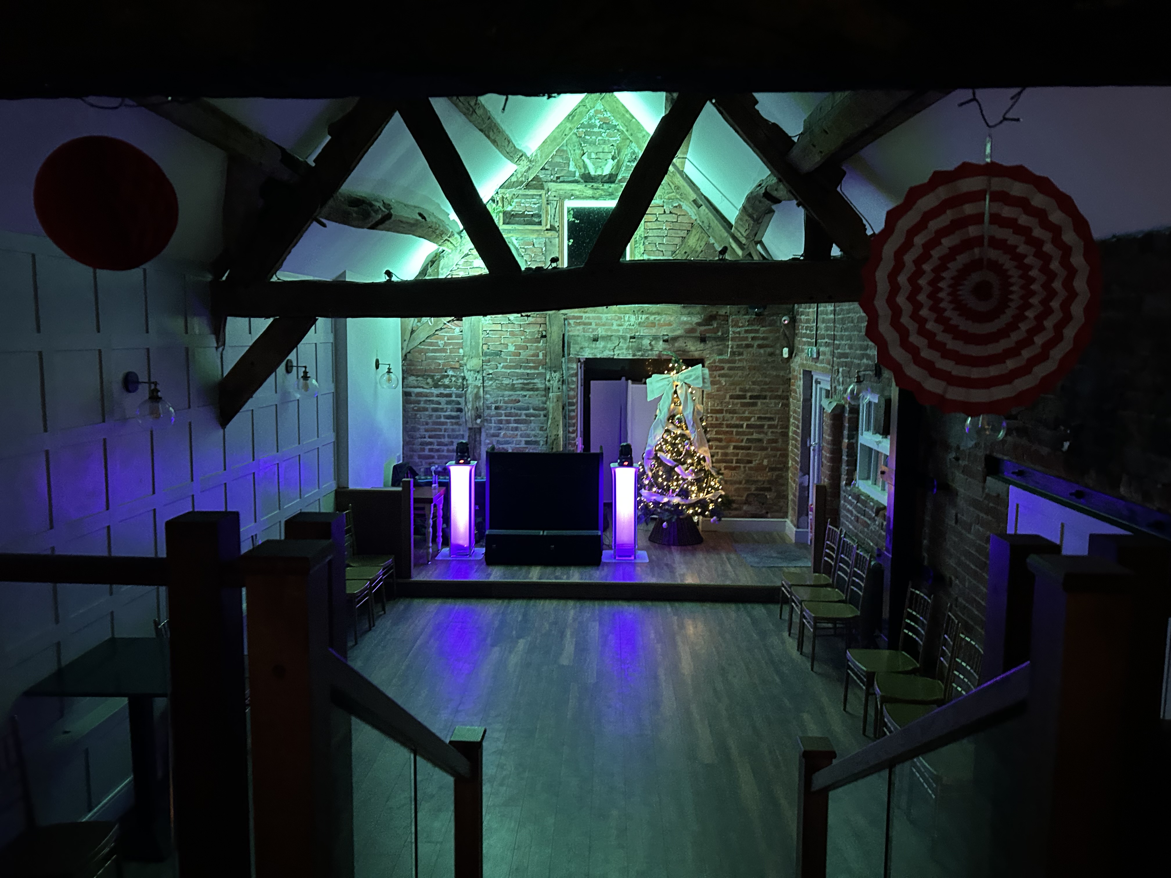 A rustic venue features exposed wooden beams and a warm wooden floor, complemented by vibrant lighting and a decorated Christmas tree, creating a festive atmosphere.
