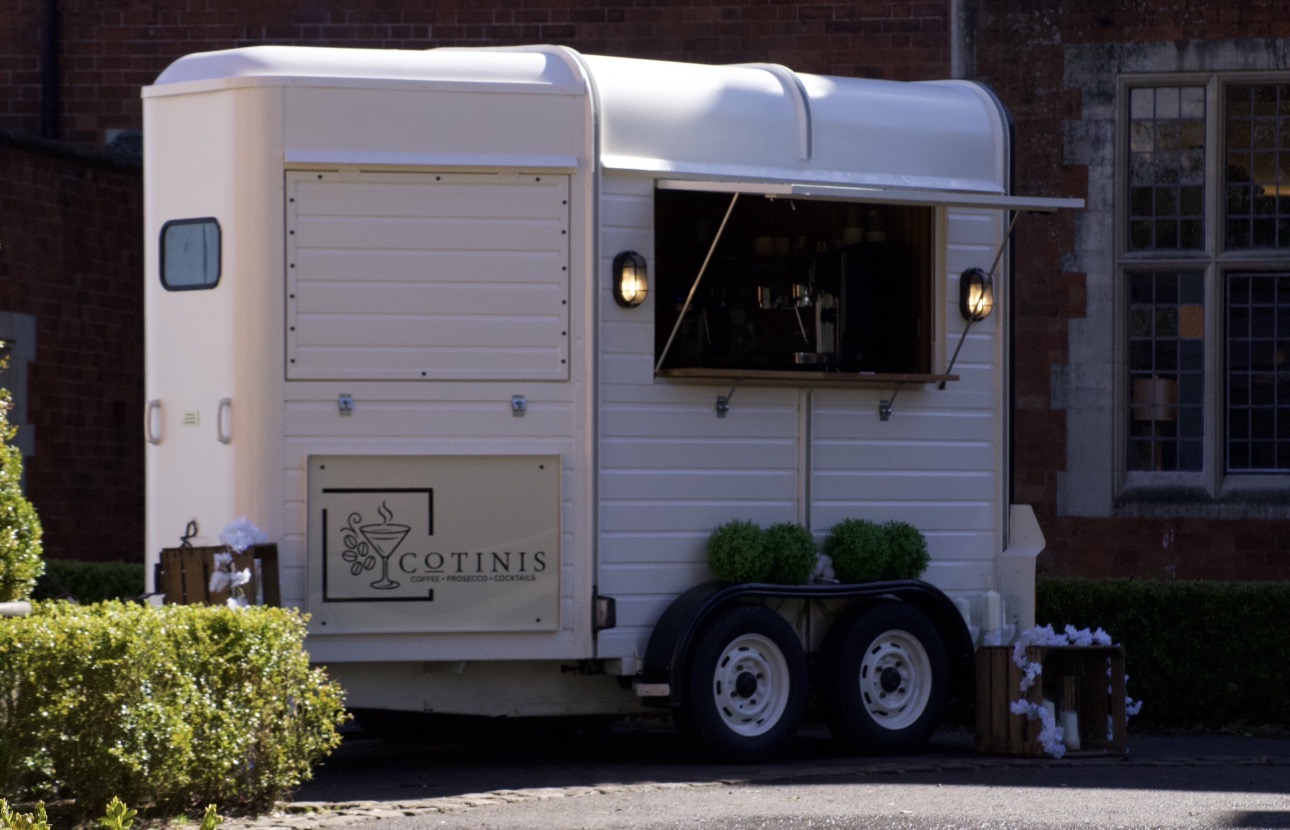 A vintage-style mobile bar with a white exterior, featuring a wooden serving window, is surrounded by lush greenery, creating a charming atmosphere.