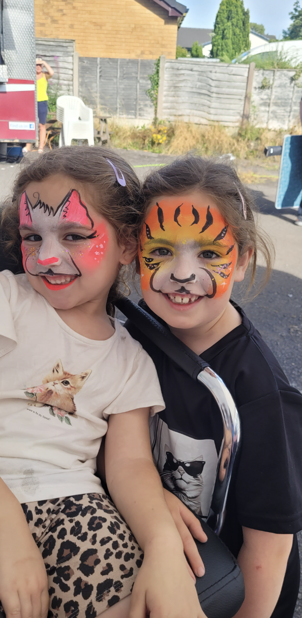 Two smiling children with tiger face paint sit on a stroller at an outdoor party, bright daylight, colorful catering atmosphere.
