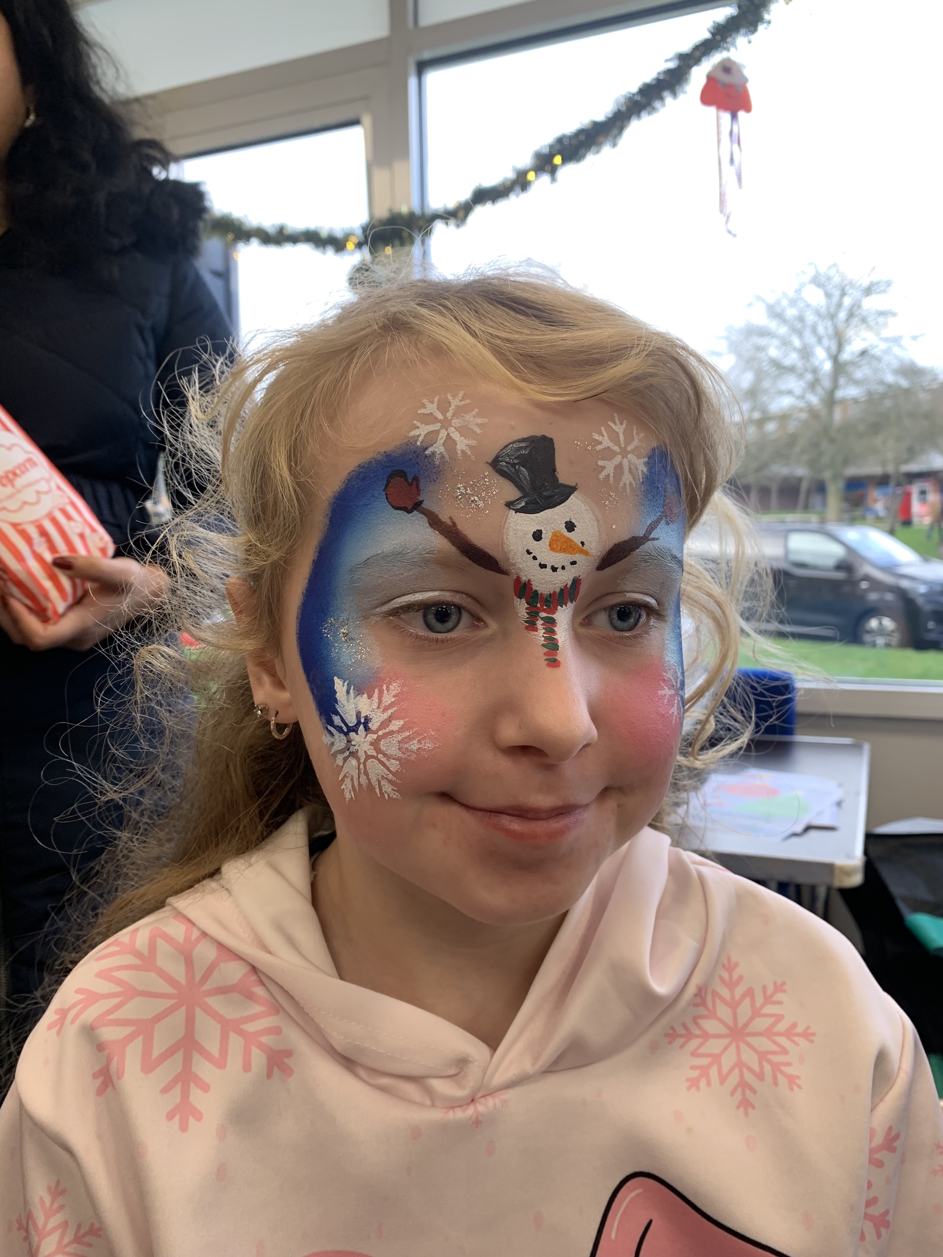 A girl with snowflake face paint, featuring a colorful snowman design, smiles softly. The cozy, festive atmosphere is enhanced by natural light.