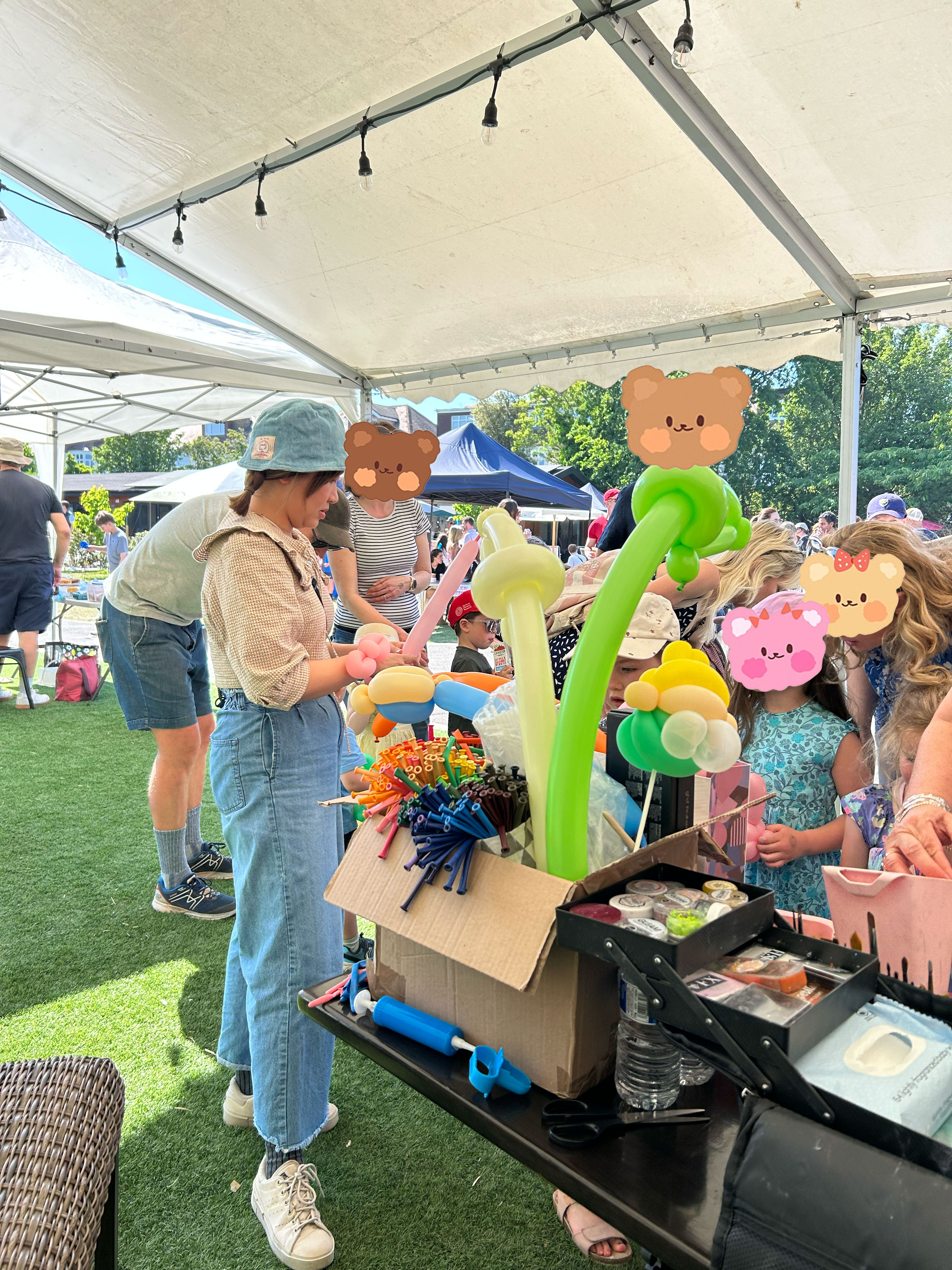 A vibrant outdoor scene features a balloon artist engaging with children under a tent, colorful balloons and supplies spread across a table, enhancing the festive atmosphere.