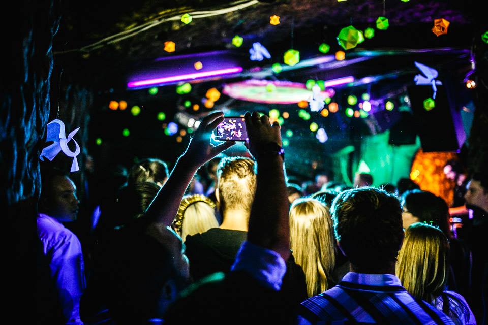 A lively crowd enjoys a vibrant nightclub, illuminated by purple and green lights, with geometric decor and a high-energy atmosphere.