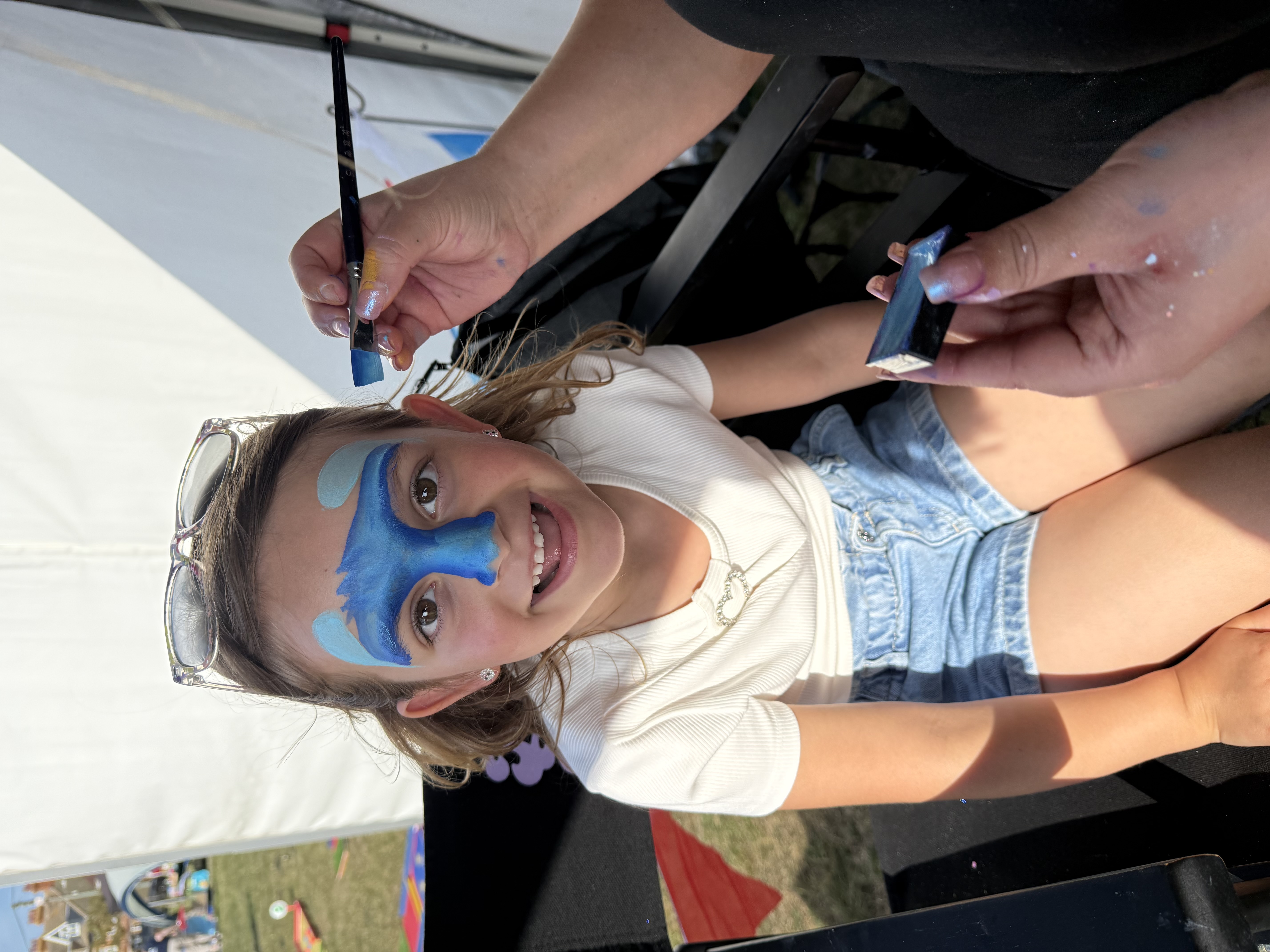 Smiling child at outdoor face-painting booth, blue butterfly design applied with brushes, black chair, bright daylight, clean event catering vibe.