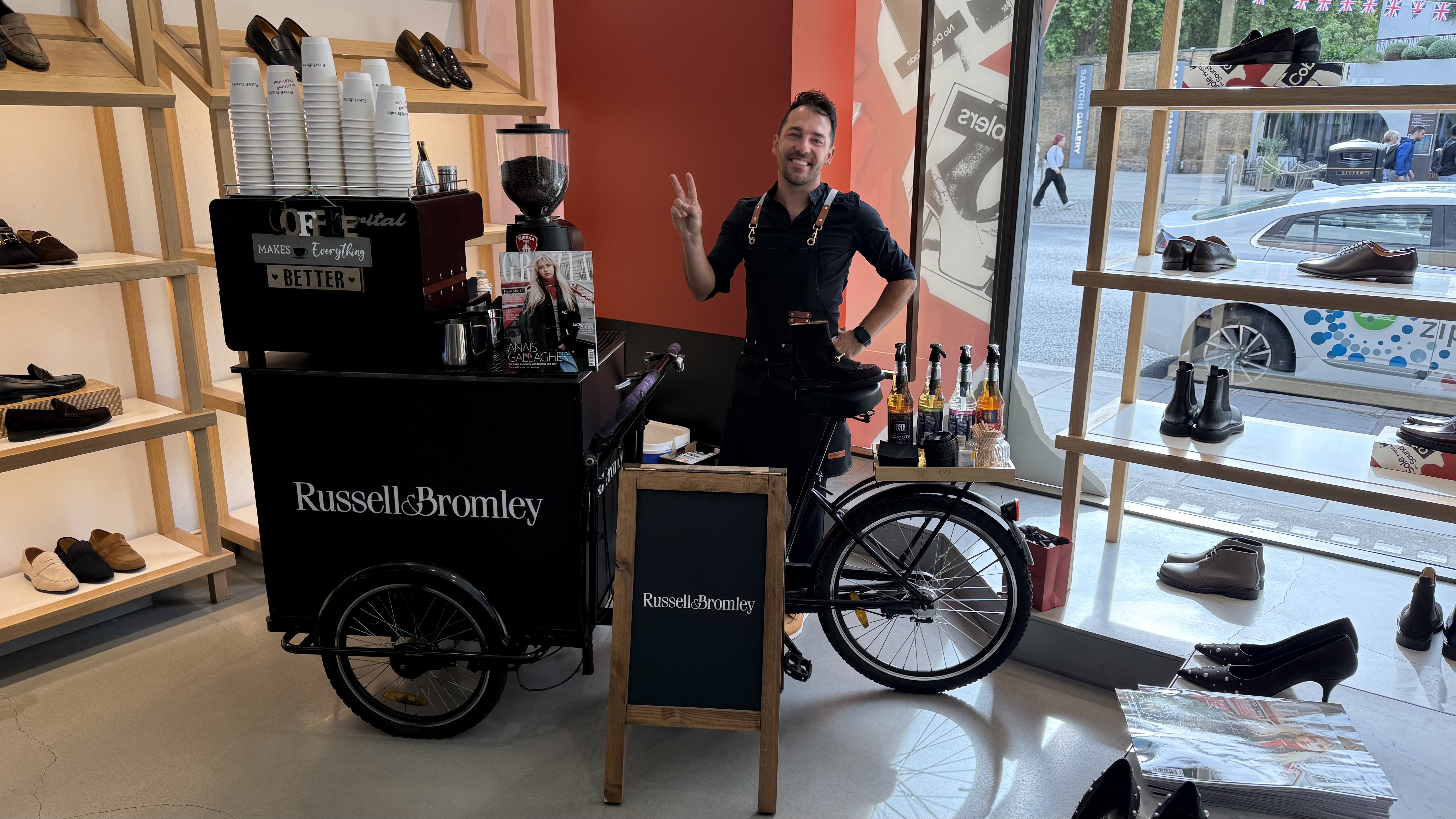 A stylish shoe store features a barista on a vintage bike cart, serving coffee amidst wooden shelves displaying elegant footwear and magazines.