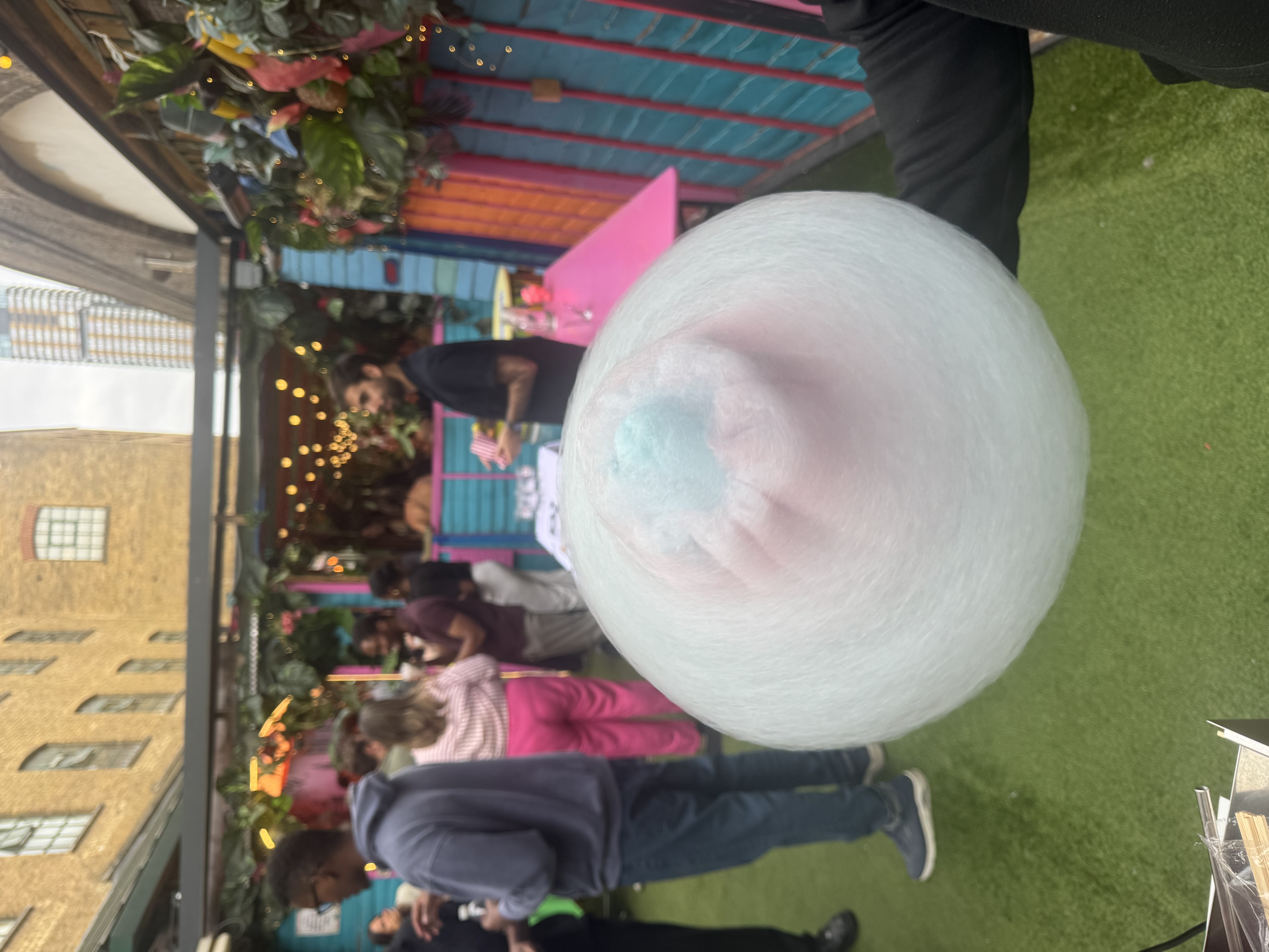 A vibrant outdoor space features a cotton candy swirl in pastel pink and blue, surrounded by lush greenery and colorful decor, evoking cheerful festive vibes.