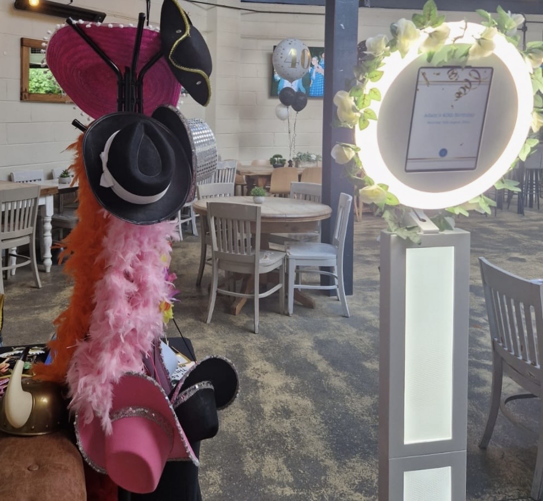 A whimsical birthday setup features a glowing photo booth surrounded by colorful hats and a feather boa on a cozy leather sofa, enhancing the celebratory vibe.