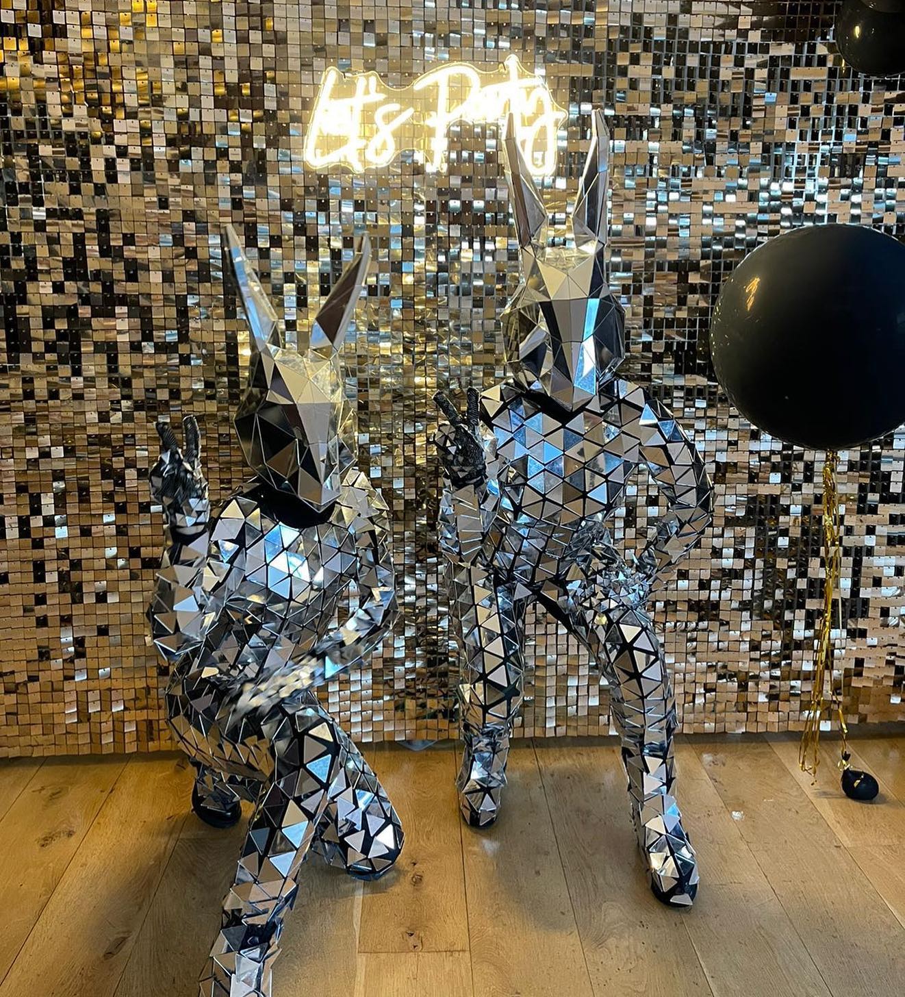 Two mirrored bunny figures pose playfully in front of a glimmering gold backdrop with a neon "Let's Party" sign, enhancing a festive vibe.