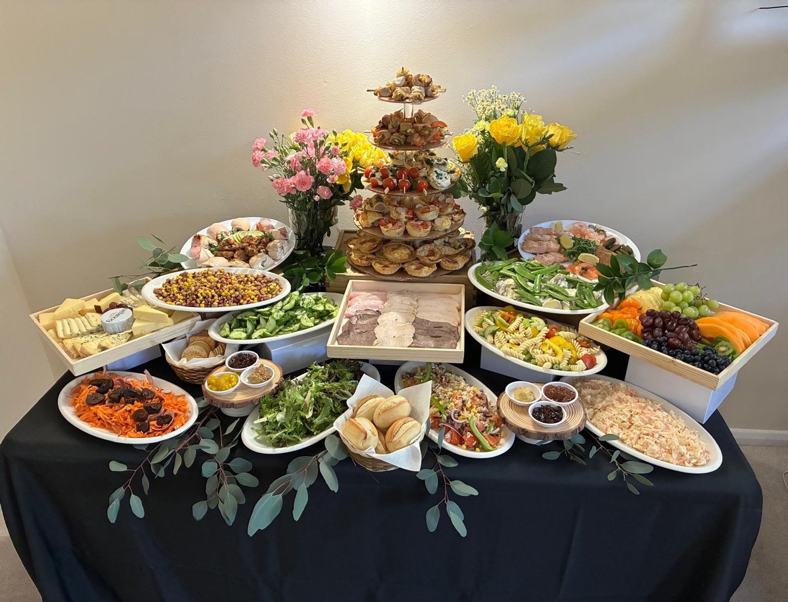 Elegant catering buffet on a black-draped table, with tiered finger sandwiches, salads, fruit, pastries, and polished glassware under warm light.