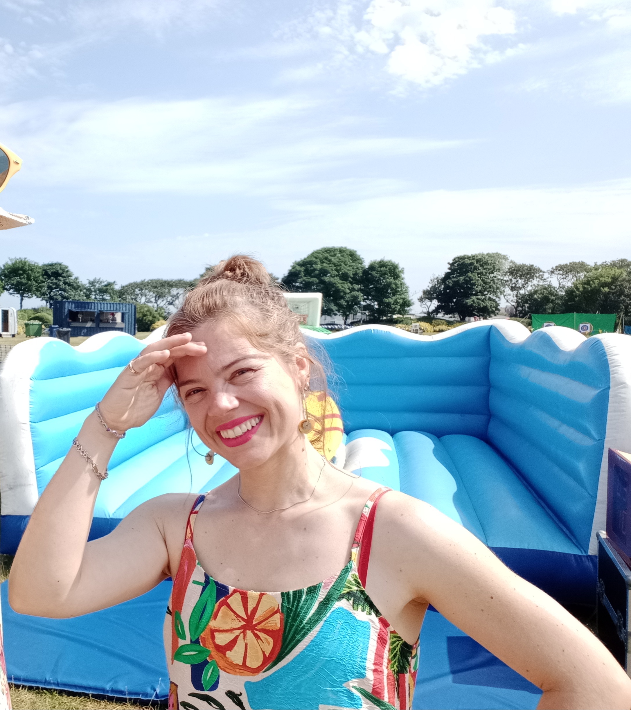 Smiling woman in a tropical-print dress poses by a blue-and-white inflatable sofa outdoors, under bright daylight at a relaxed event.