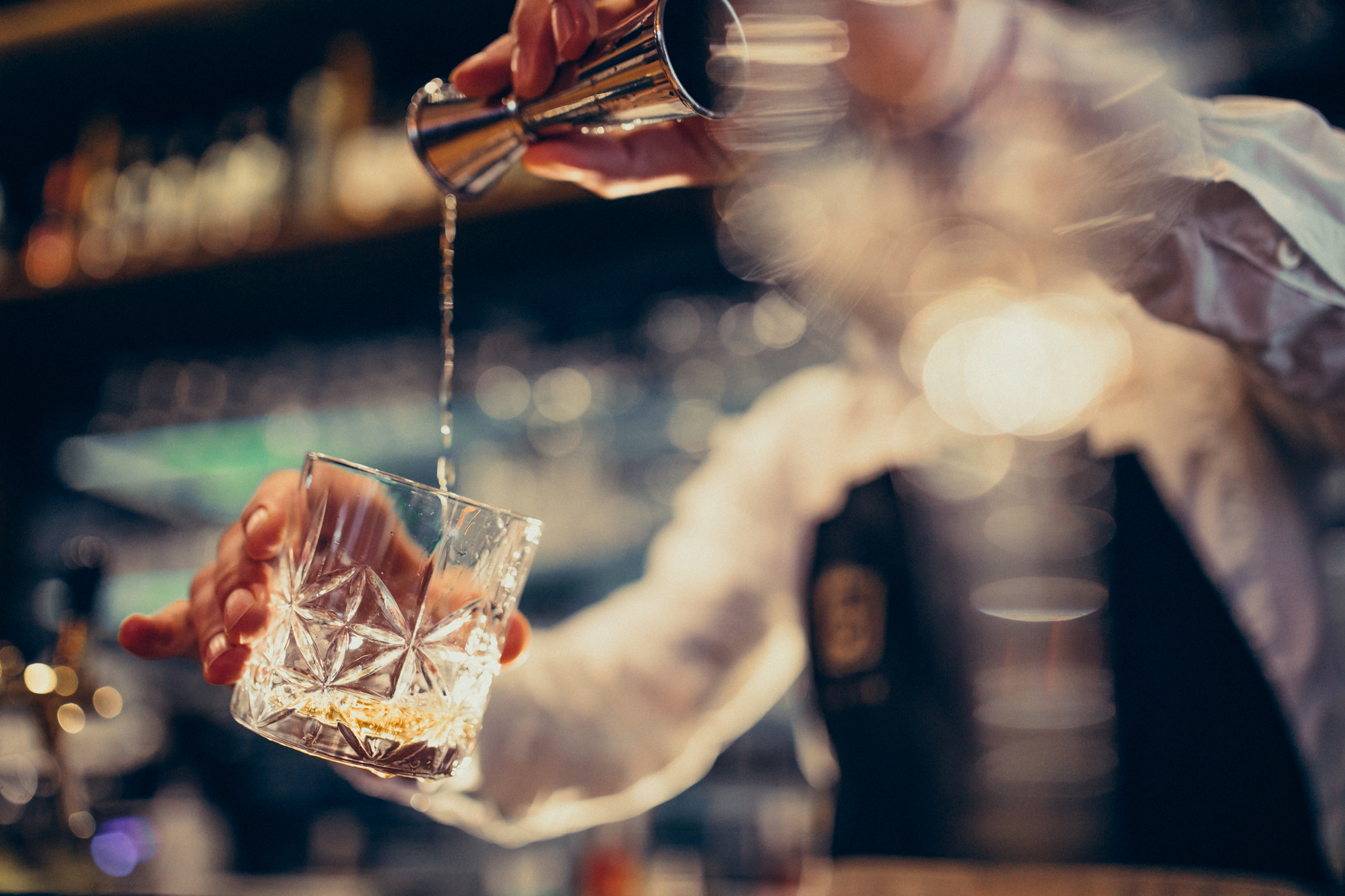 A bartender skillfully pours a golden spirit into a crystal tumbler, surrounded by blurred bar details, creating a cozy, elegant vibe.
