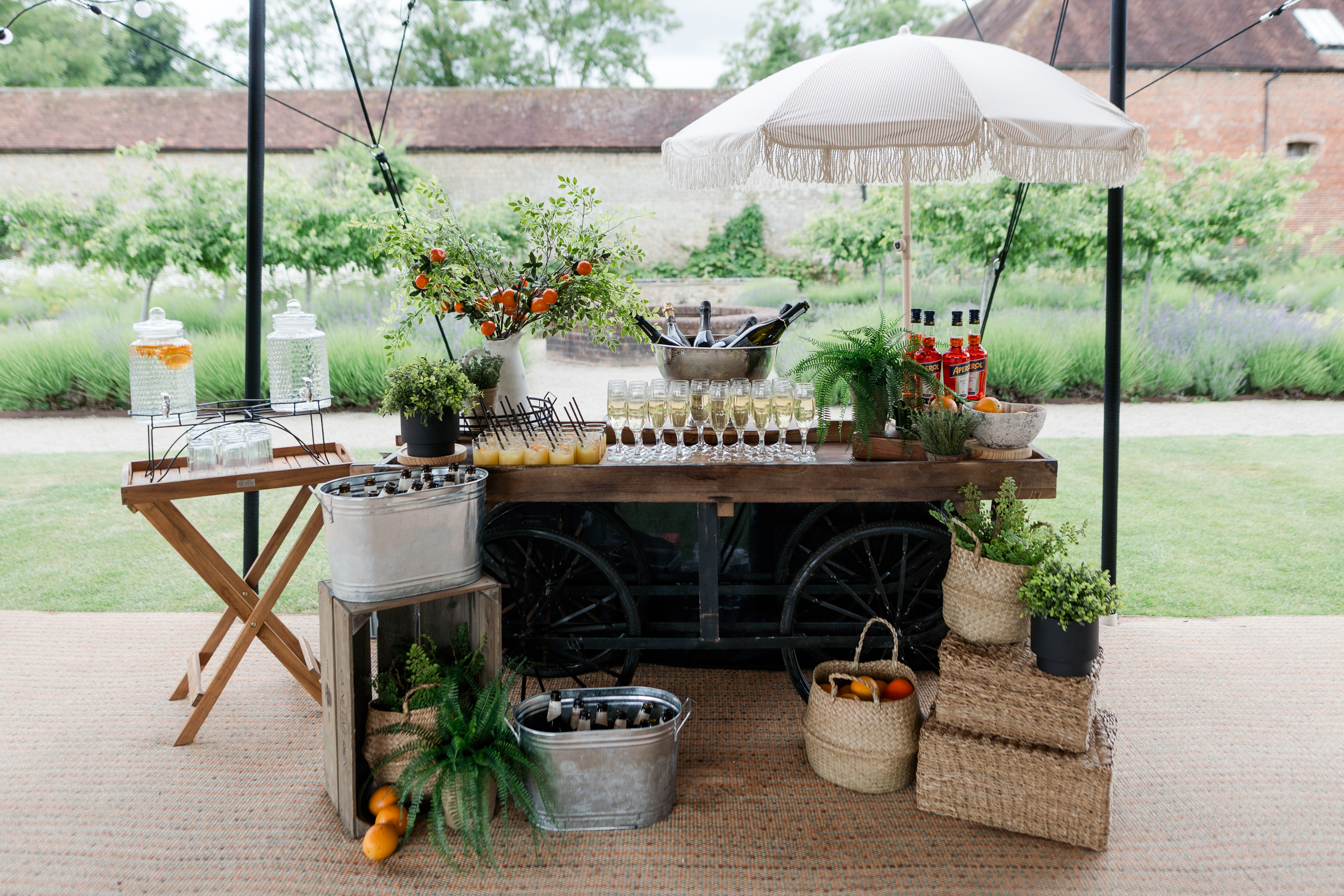 A rustic wooden catering cart displays sparkling water dispensers, vibrant greenery, and elegant glassware under a chic striped umbrella, evoking a relaxed, inviting atmosphere.