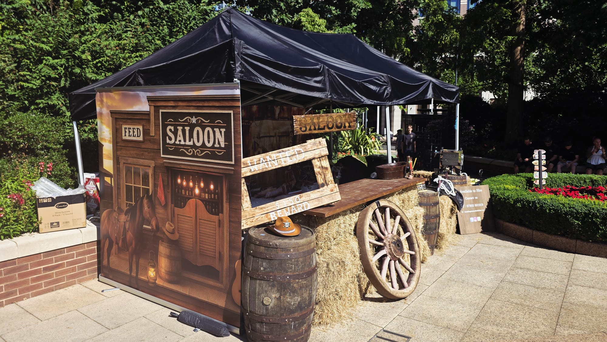 A rustic "saloon" booth features wooden barrels and hay bales under a black tent, surrounded by vibrant flowers in bright daylight.