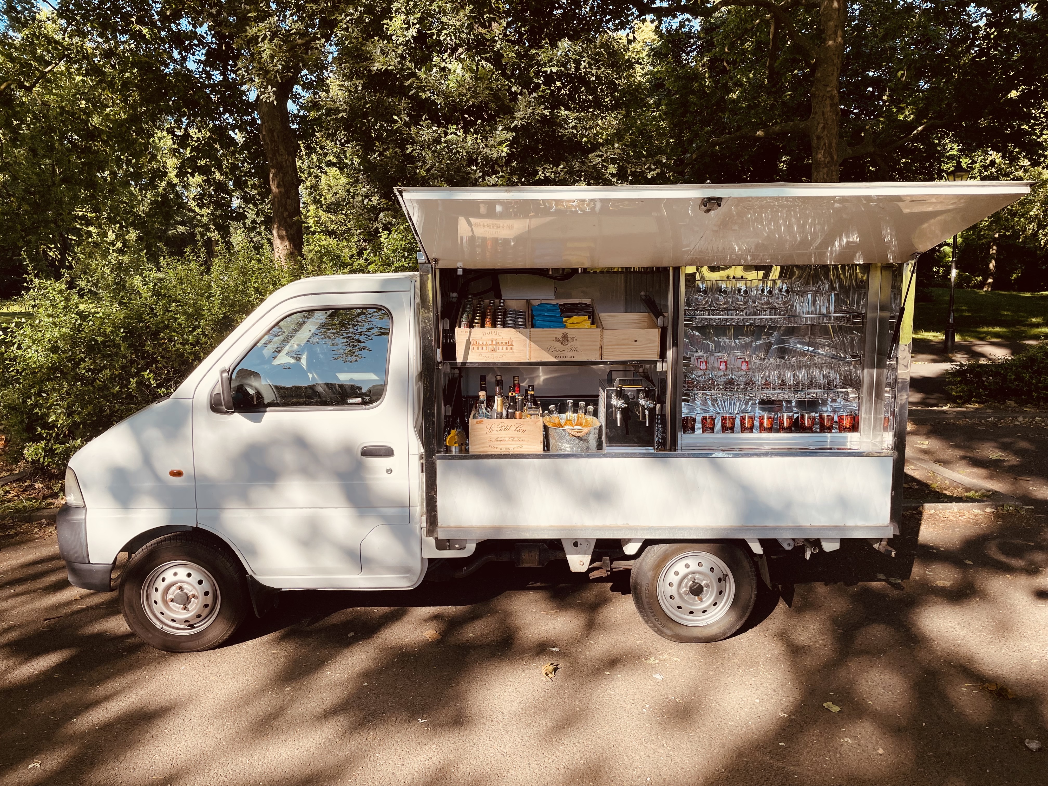 A vintage-style catering van showcases neatly arranged glassware and beverages under natural sunlight, highlighting its polished elegance.