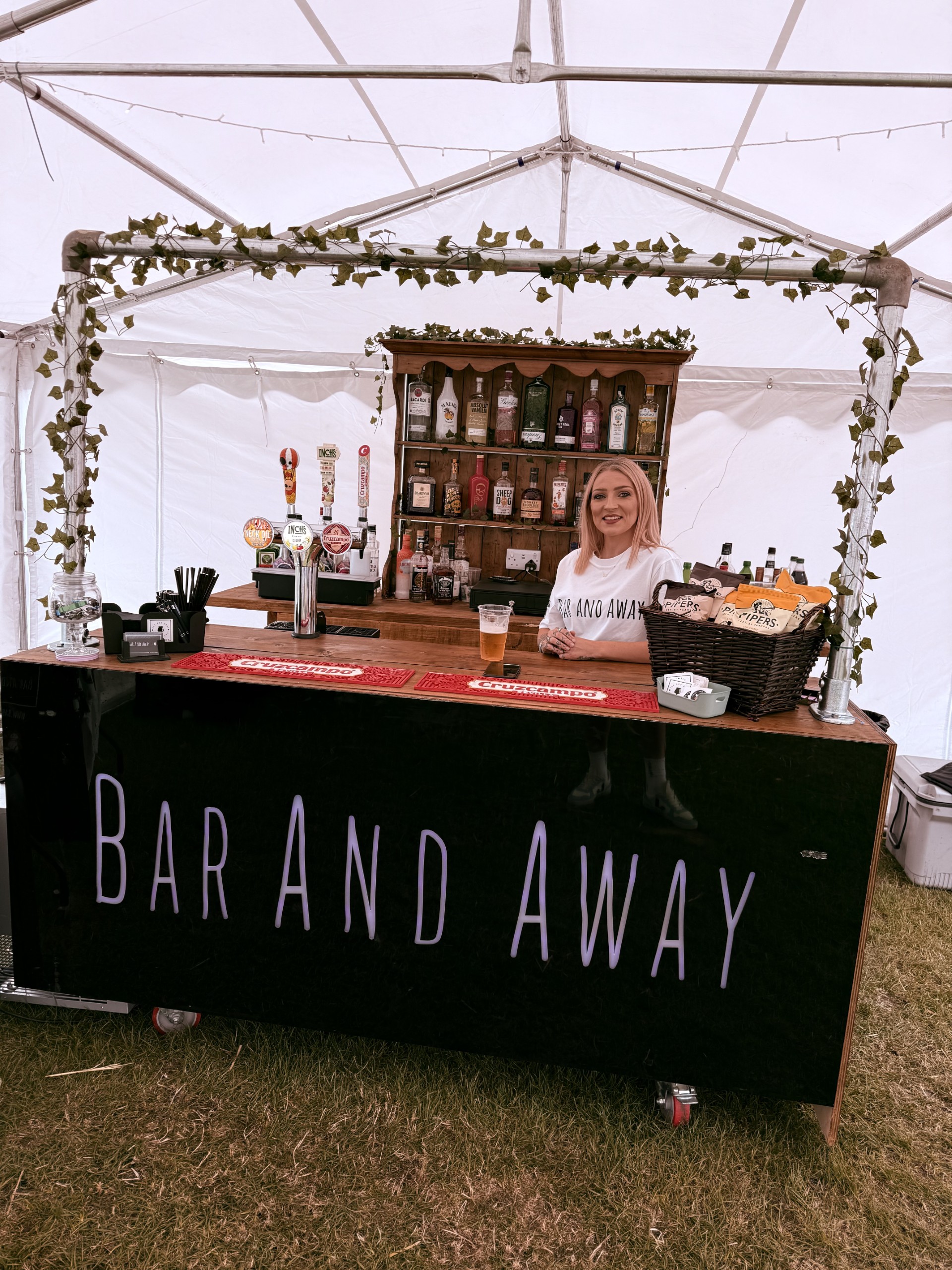 A stylish bar setup under a white tent features a sleek black counter displaying craft beers, liquor bottles, and a cheerful bartender amidst greenery.