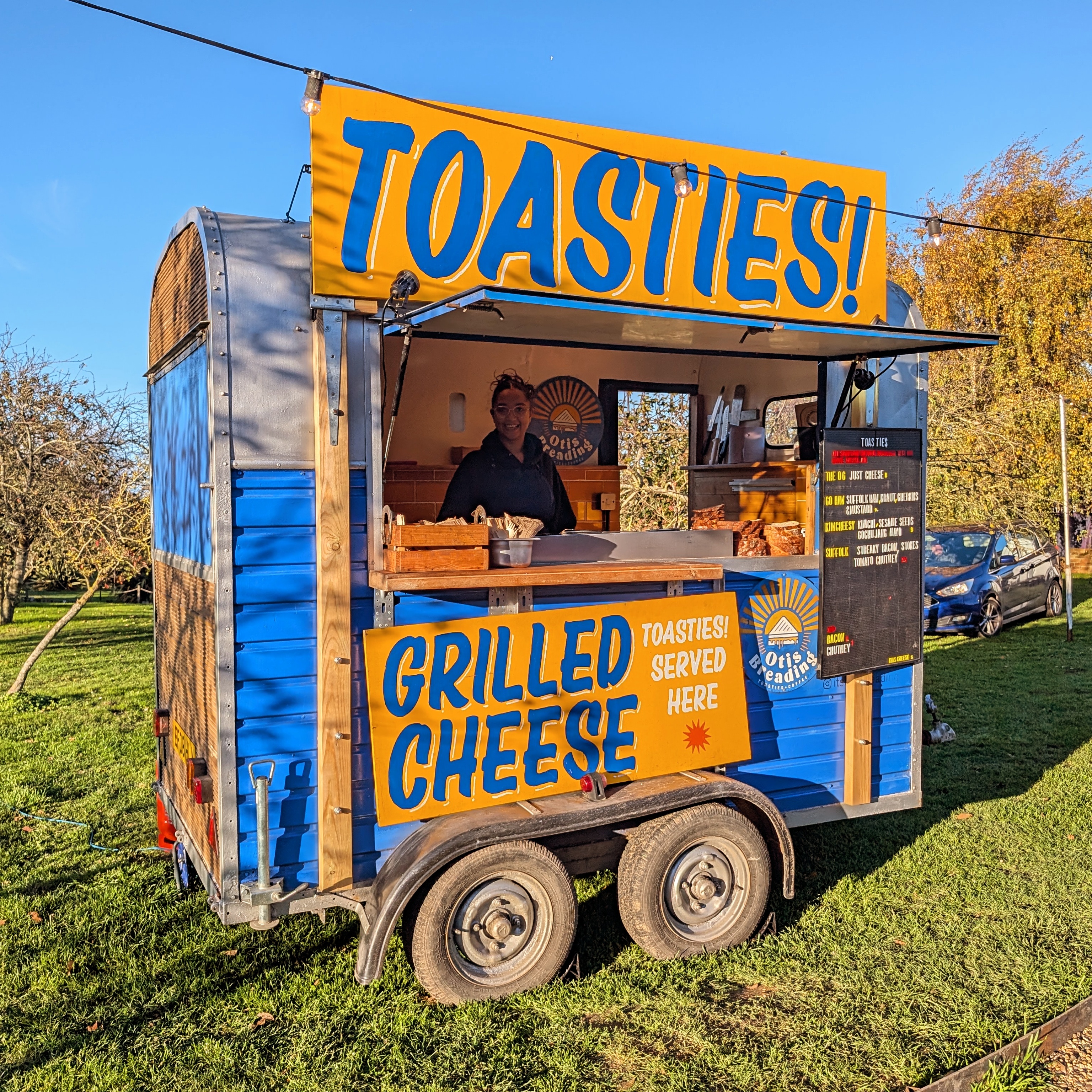 A vibrant food truck showcases "Grilled Cheese" offerings, featuring a cheerful vendor amidst sunny outdoor vibes, inviting patrons to enjoy.