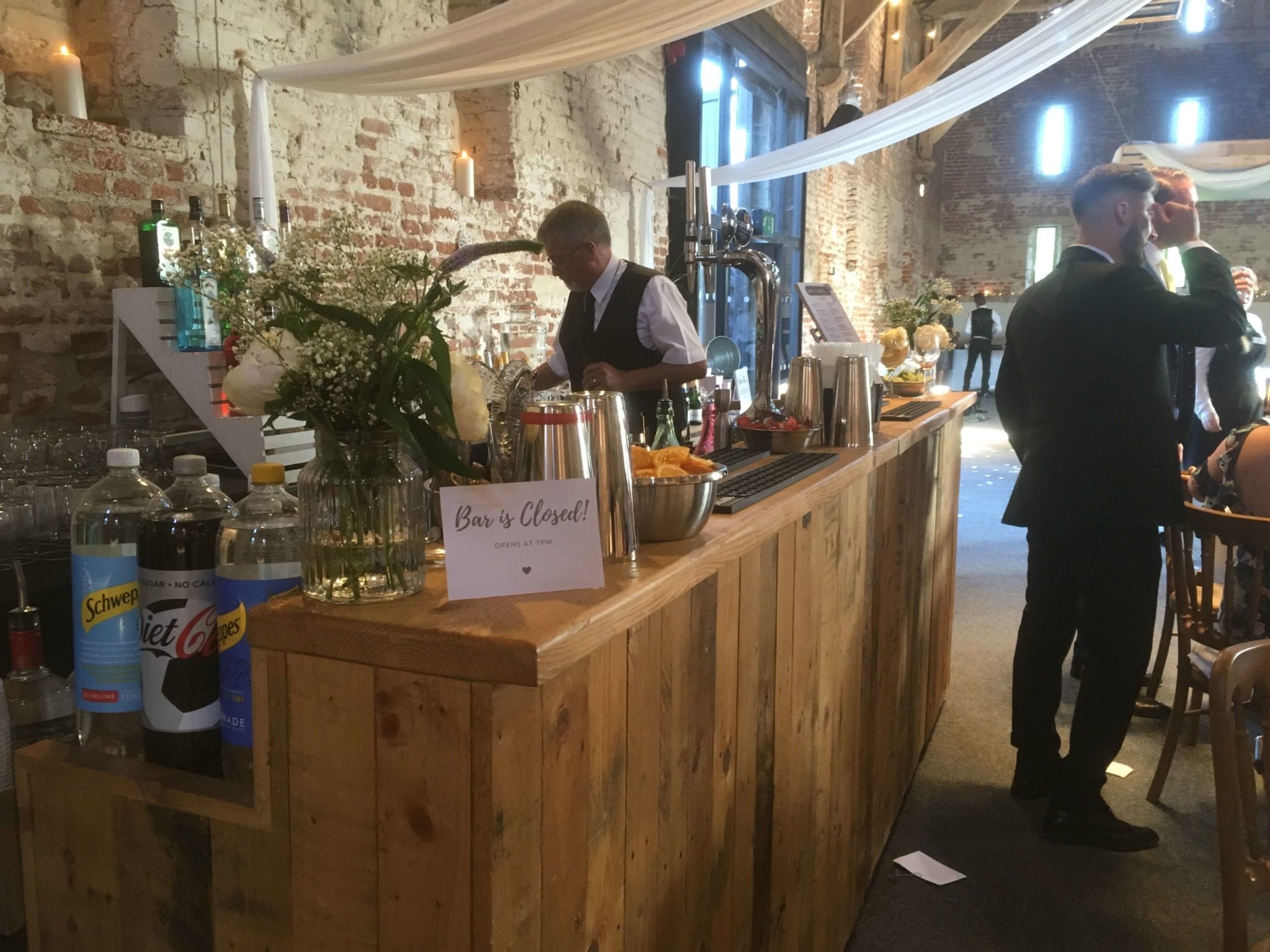 A rustic wooden bar showcases polished glassware, assorted drinks, and fresh flowers, illuminated by warm natural light, enhancing the elegant atmosphere.