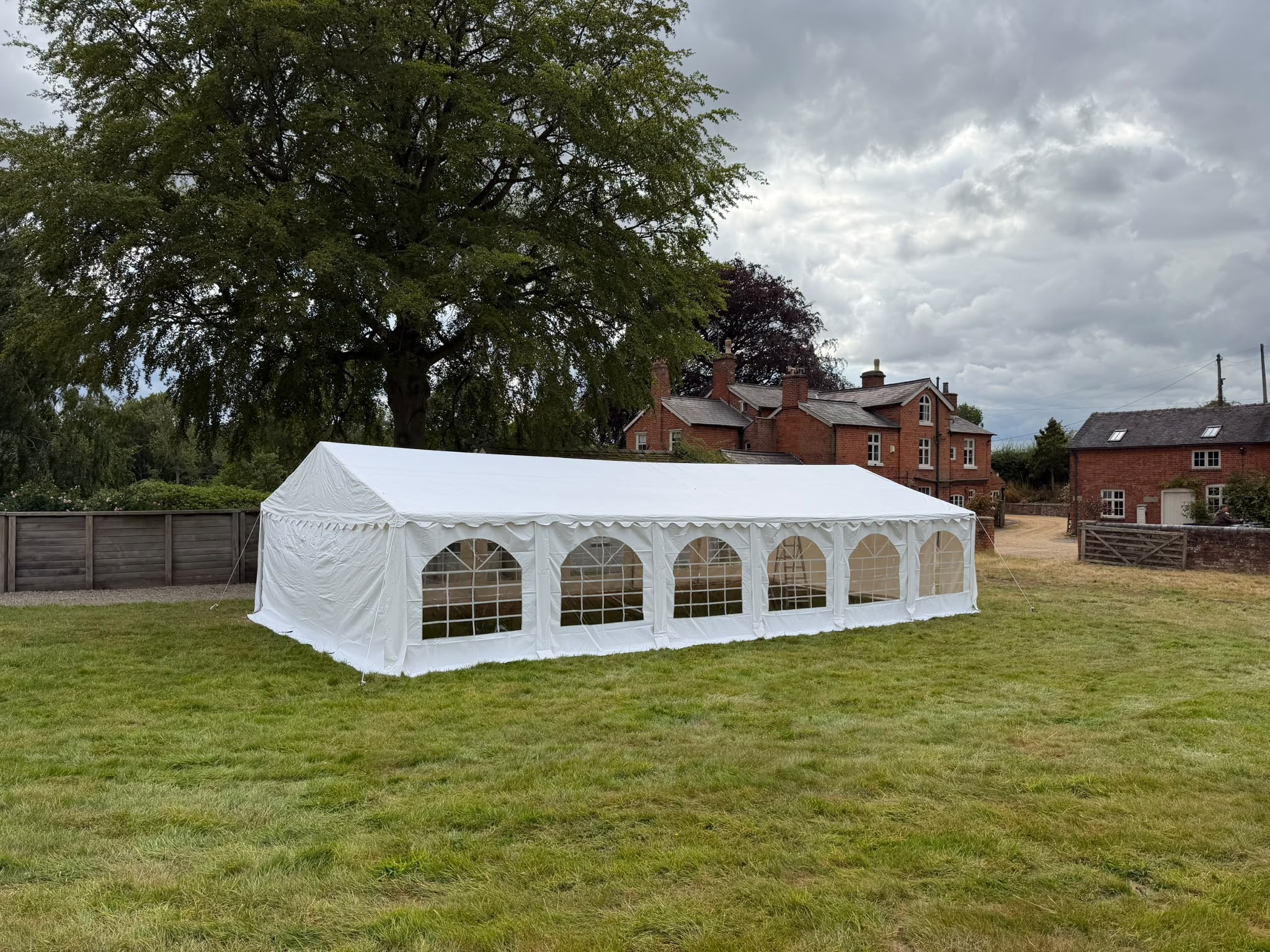 White marquee catering tent on a grassy lawn beside red-brick houses, with arched windows, polished setup, and soft overcast daylight.
