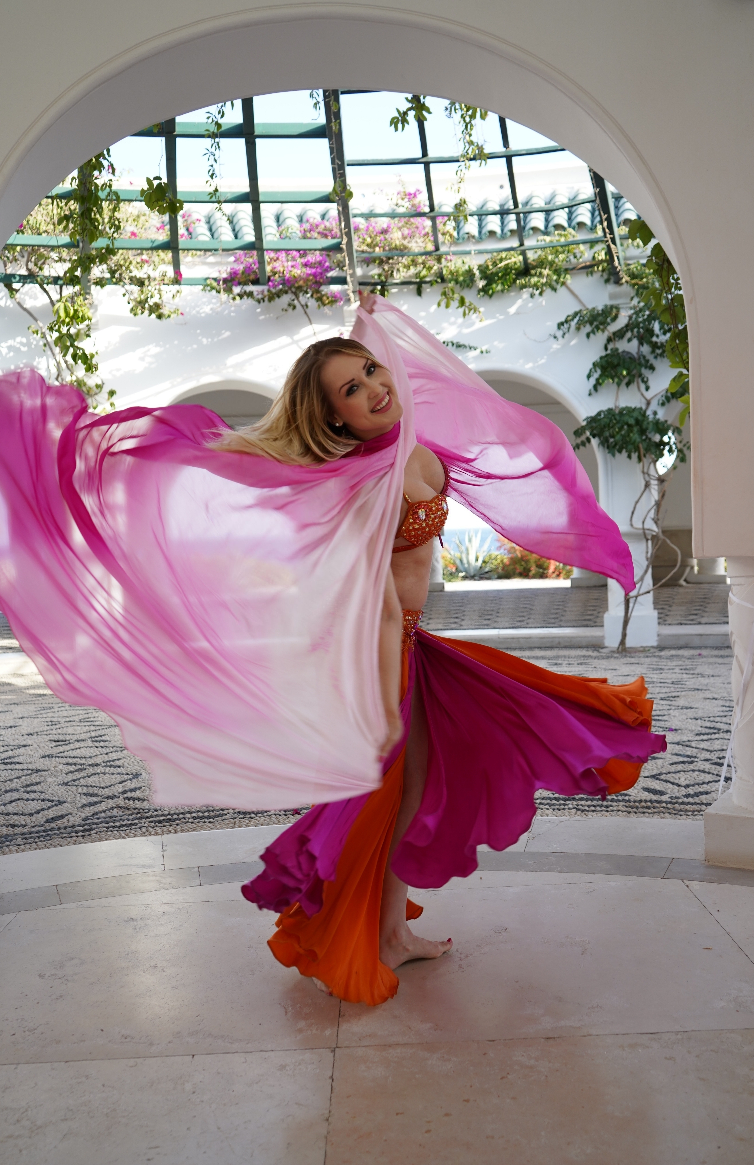 A dancer twirls gracefully in vibrant pink and orange fabric, illuminated by soft daylight amidst a lush, elegant backdrop, showcasing artful movement.