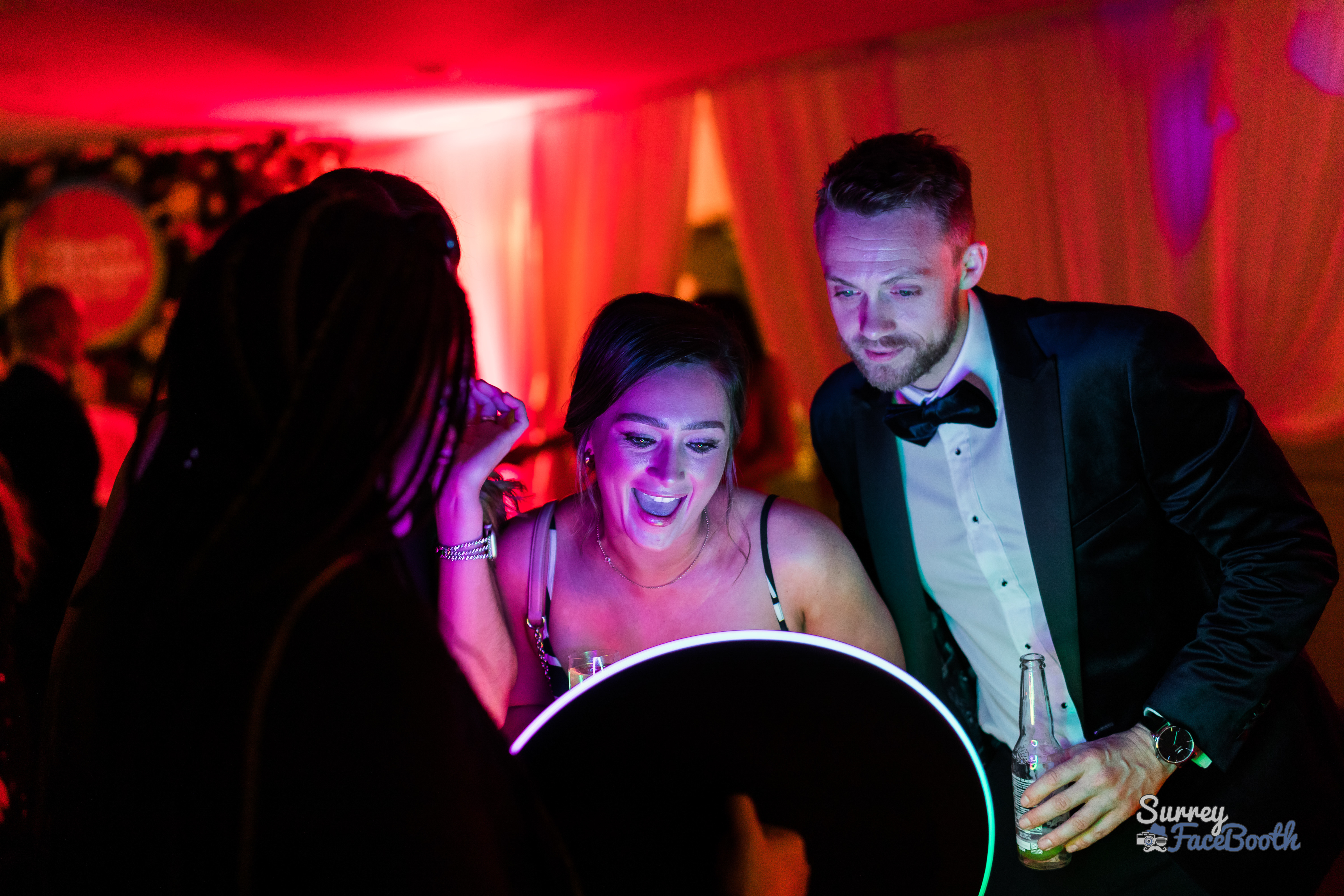 Under vibrant pink and purple lighting, a group of elegantly dressed individuals laugh and engage with a sleek photo booth, creating joyful memories.