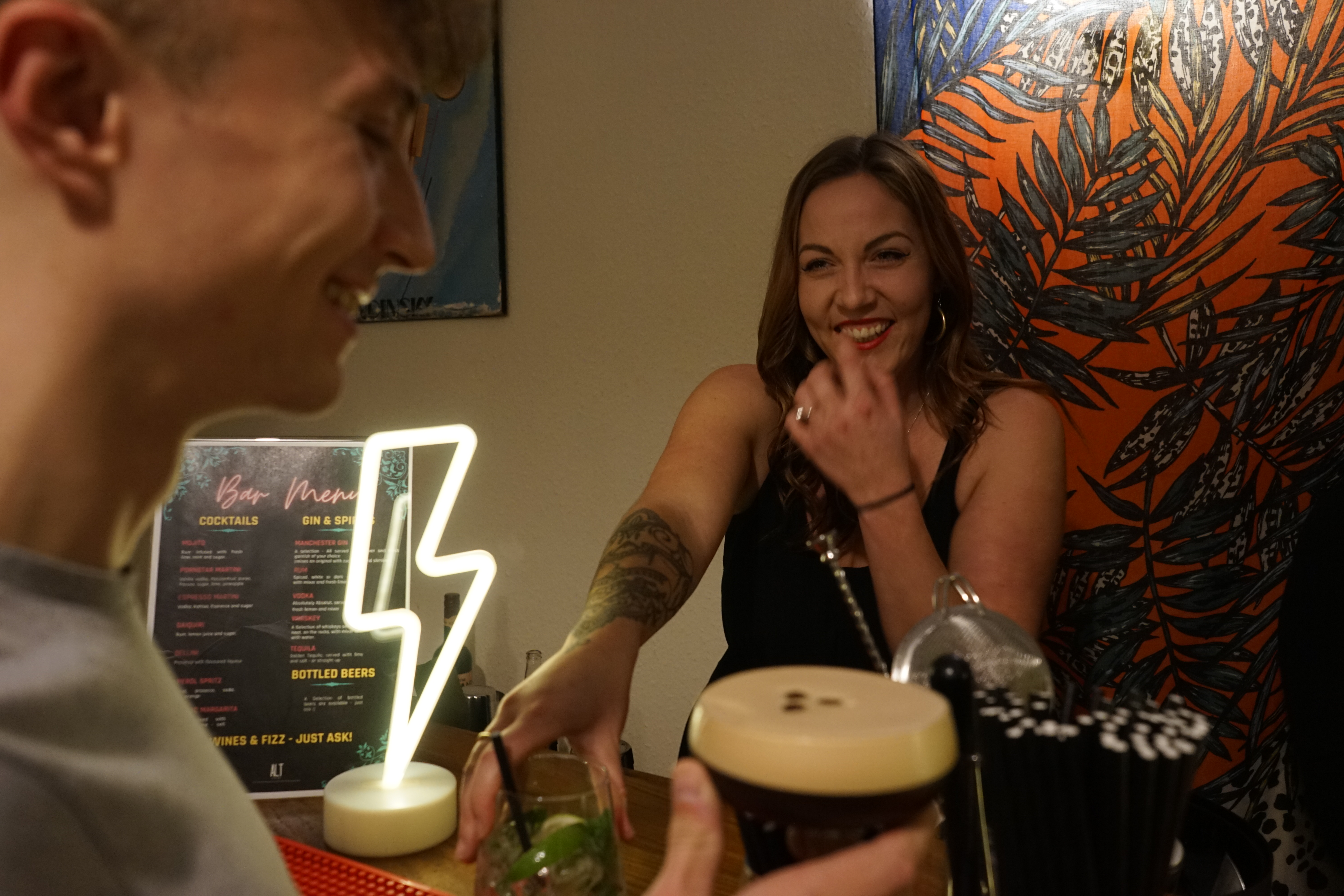 A lively bar scene features a smiling bartender handing over a cocktail, illuminated by a neon lightning bolt, with a vibrant palm-patterned backdrop.