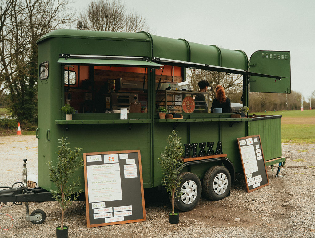 A vibrant green pizza van features a rustic wooden serving counter and cozy potted plants, offering artisanal slices in a relaxed outdoor setting.