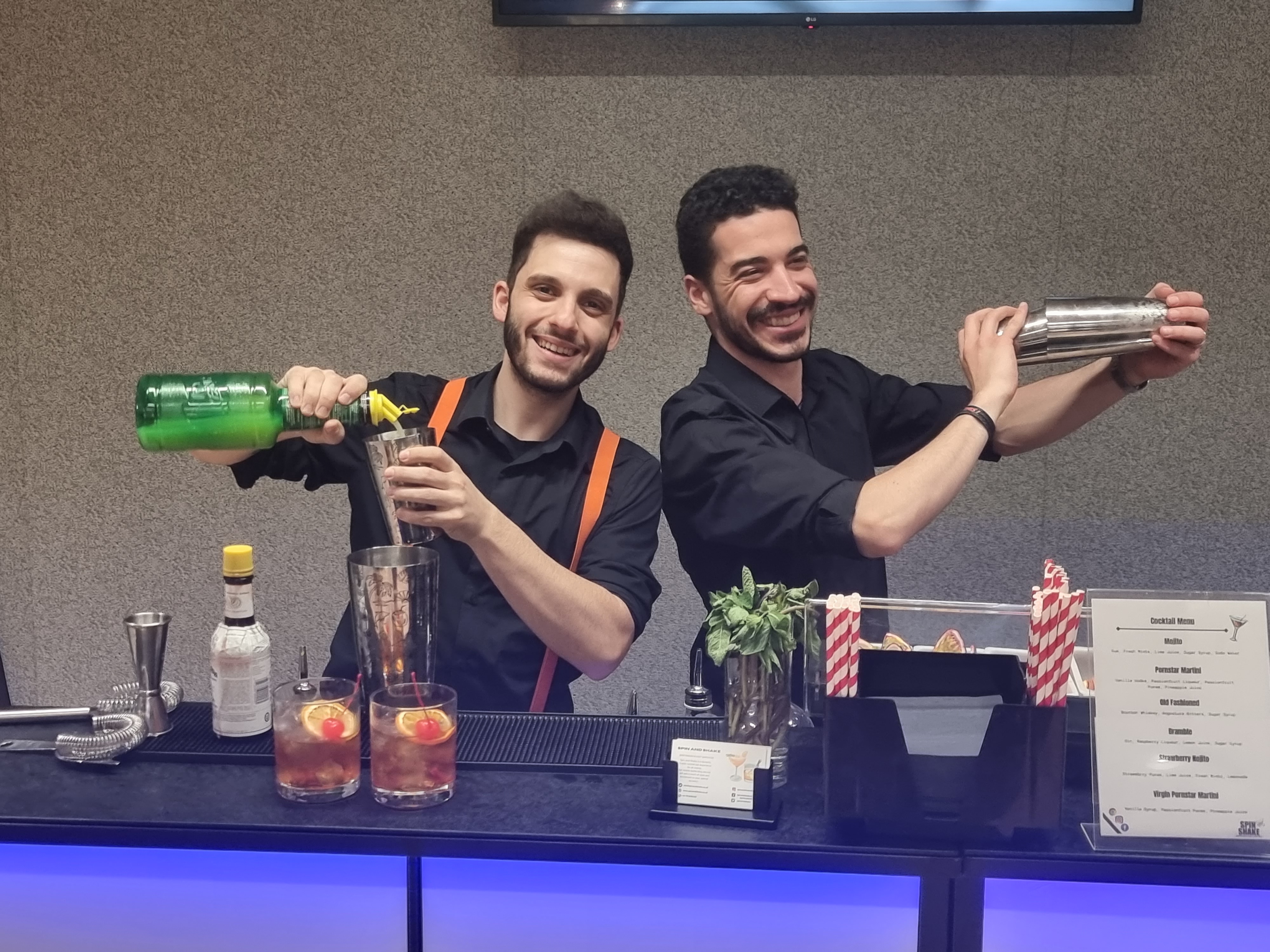 Two bartenders in black shirts with orange suspenders shake cocktail shakers at a sleek bar with colorful drinks and a menu display.