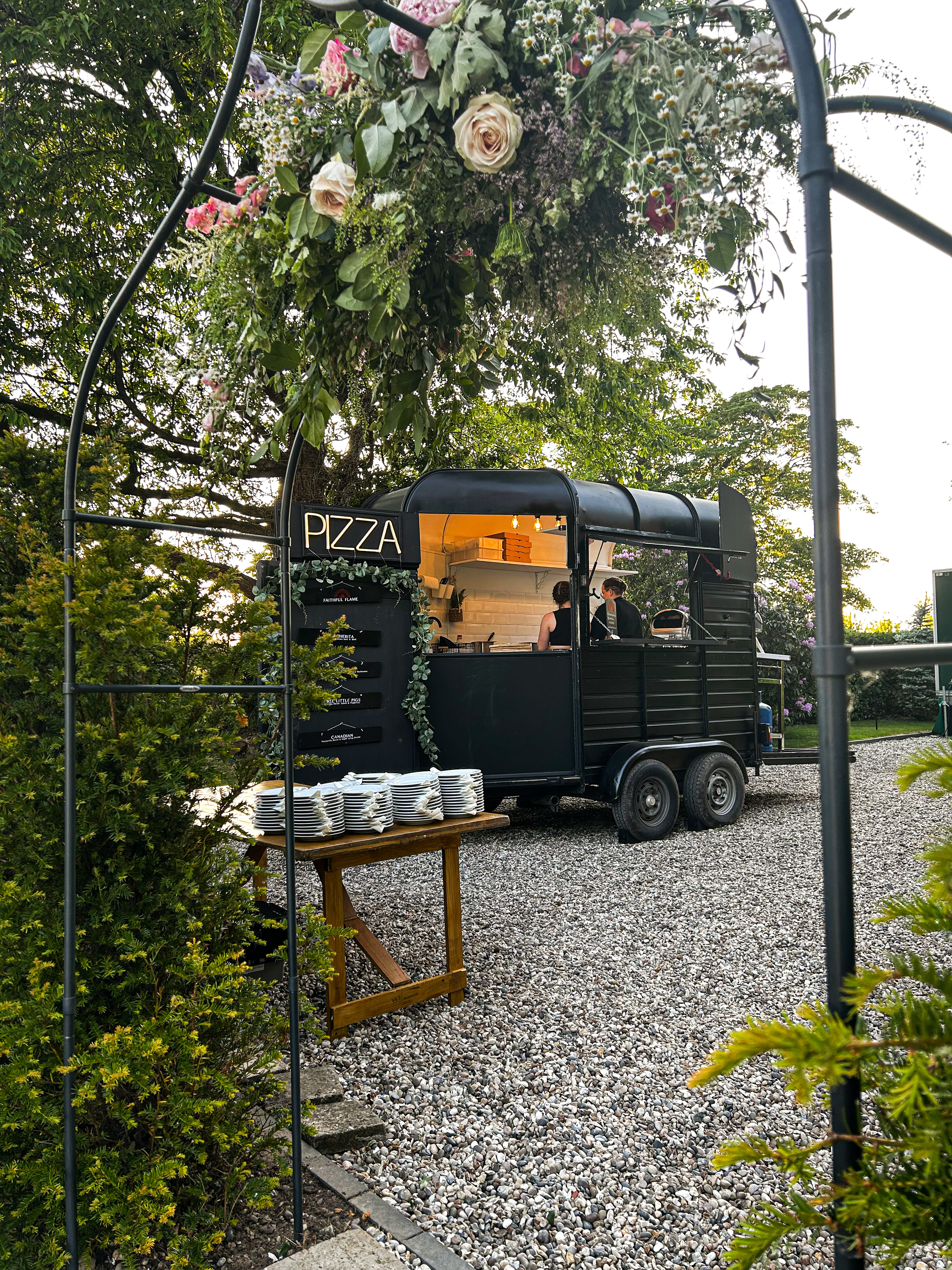 A charming mobile pizza van, surrounded by greenery, features rustic wooden tables with neatly stacked white plates, creating an inviting atmosphere.