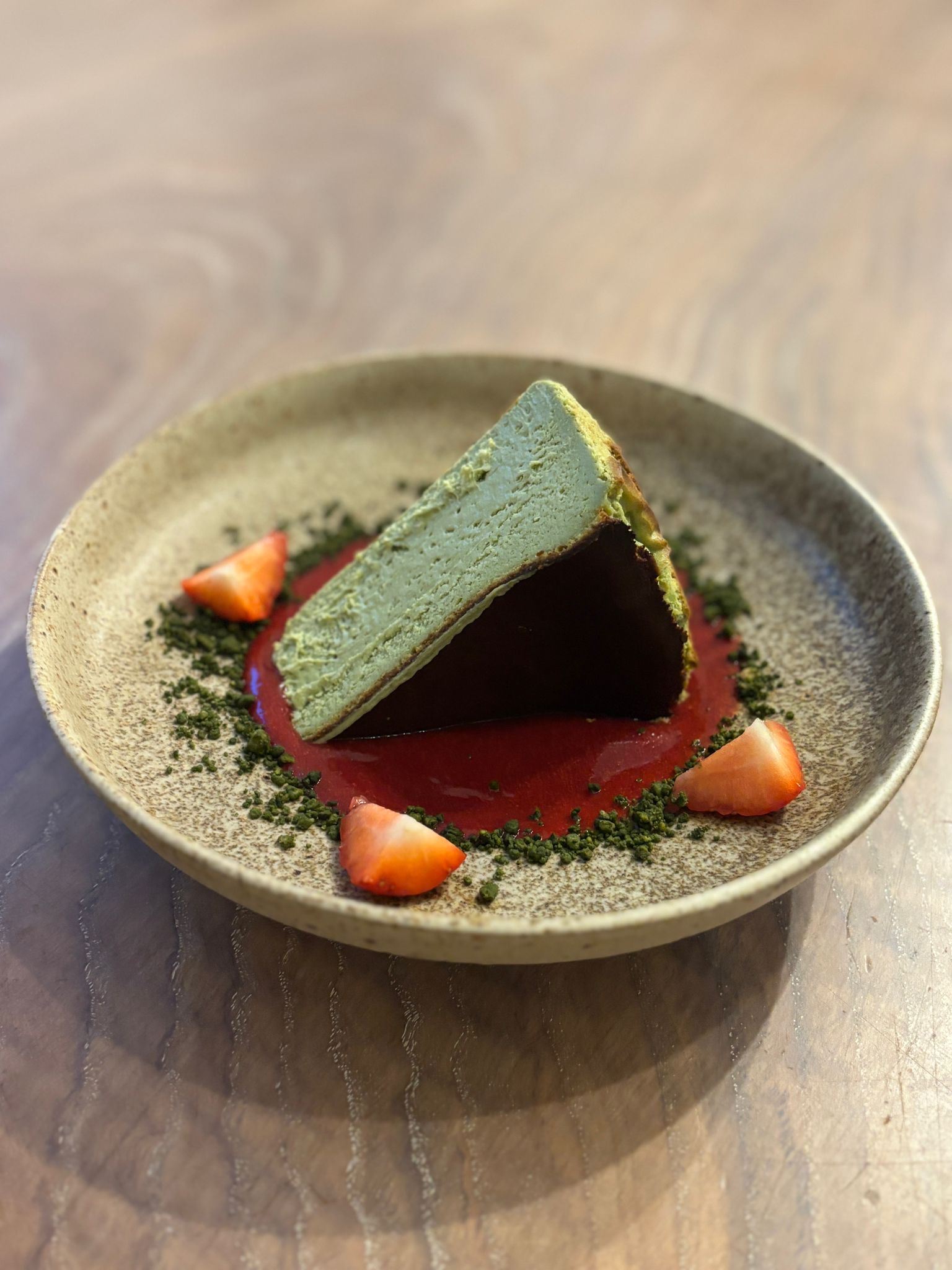 A sleek, triangular mousse dessert sits on a textured beige plate, surrounded by vibrant raspberry sauce and fresh strawberry slices, evoking elegance.