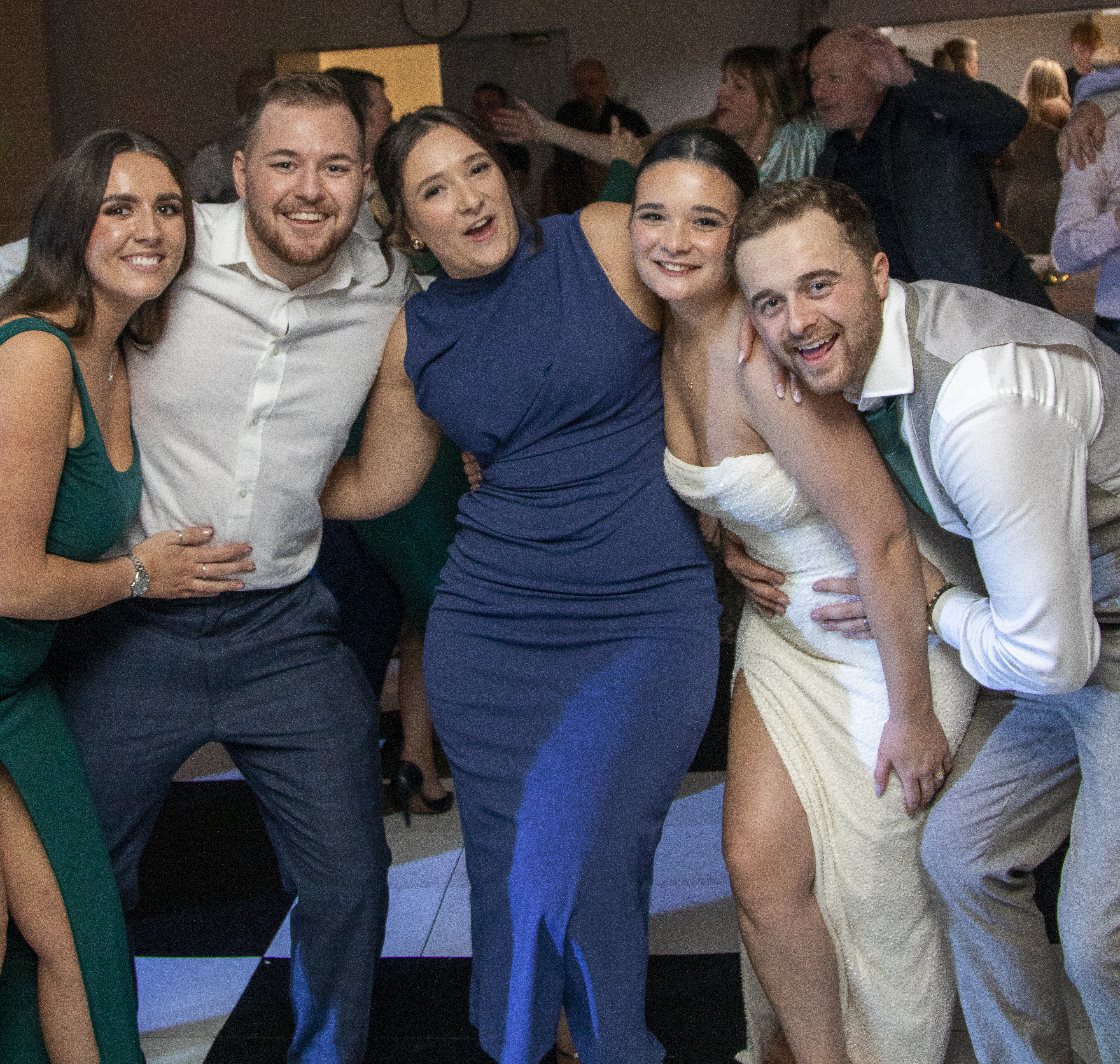 Smiling wedding guests pose in a tight group on a black-and-white dance floor, under warm reception lighting at a lively catered venue.
