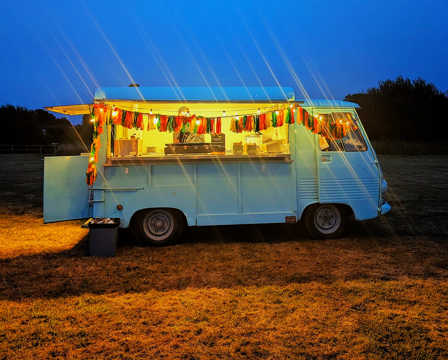 A vintage turquoise food truck glows warmly under string lights, with colorful decorations, inviting an evening catering ambiance.