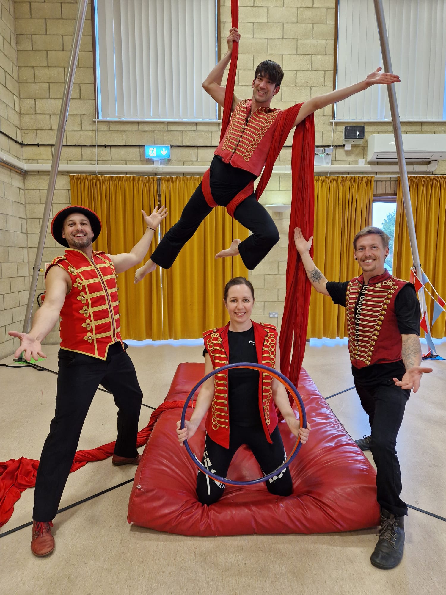 A vibrant circus scene features four performers in red-and-gold costumes, engaged in acrobatics on a padded mat, radiating excitement.