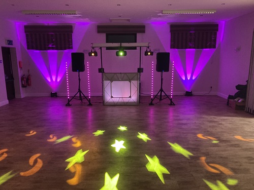 Purple-lit catering DJ booth with twin speakers, green uplights and star floor gobos on polished wood, clean modern event setup.
