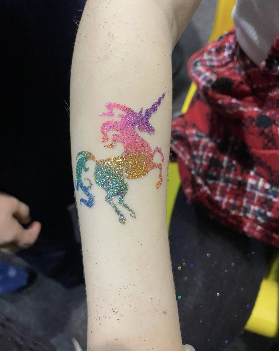 A vibrant glitter tattoo of a prancing unicorn in shades of pink, gold, and teal adorns a child's arm, shimmering in playful colors.