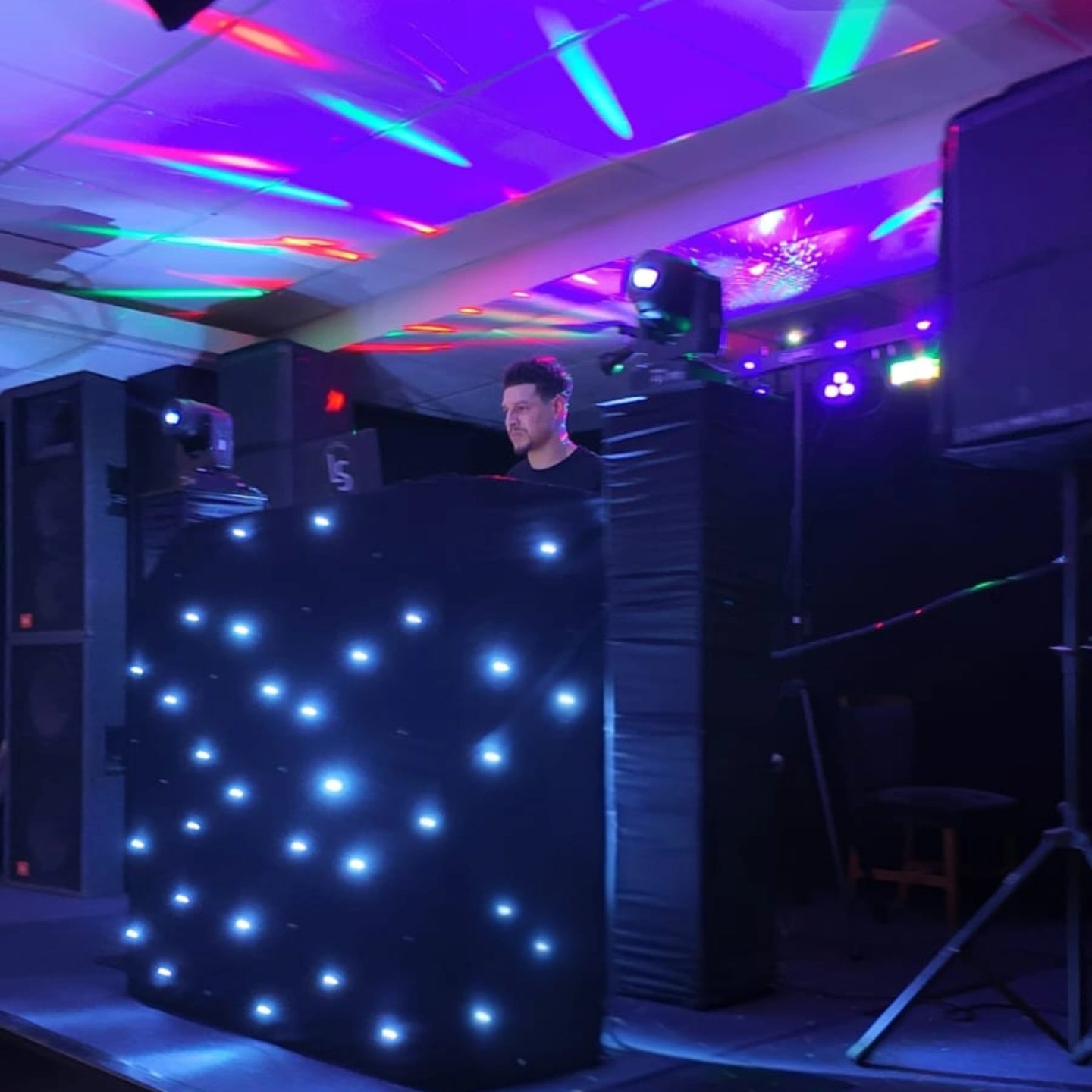 A DJ stands behind a sleek black booth, flanked by two speakers, with a festive Christmas tree adorned in blue and silver, under warm pendant lights.