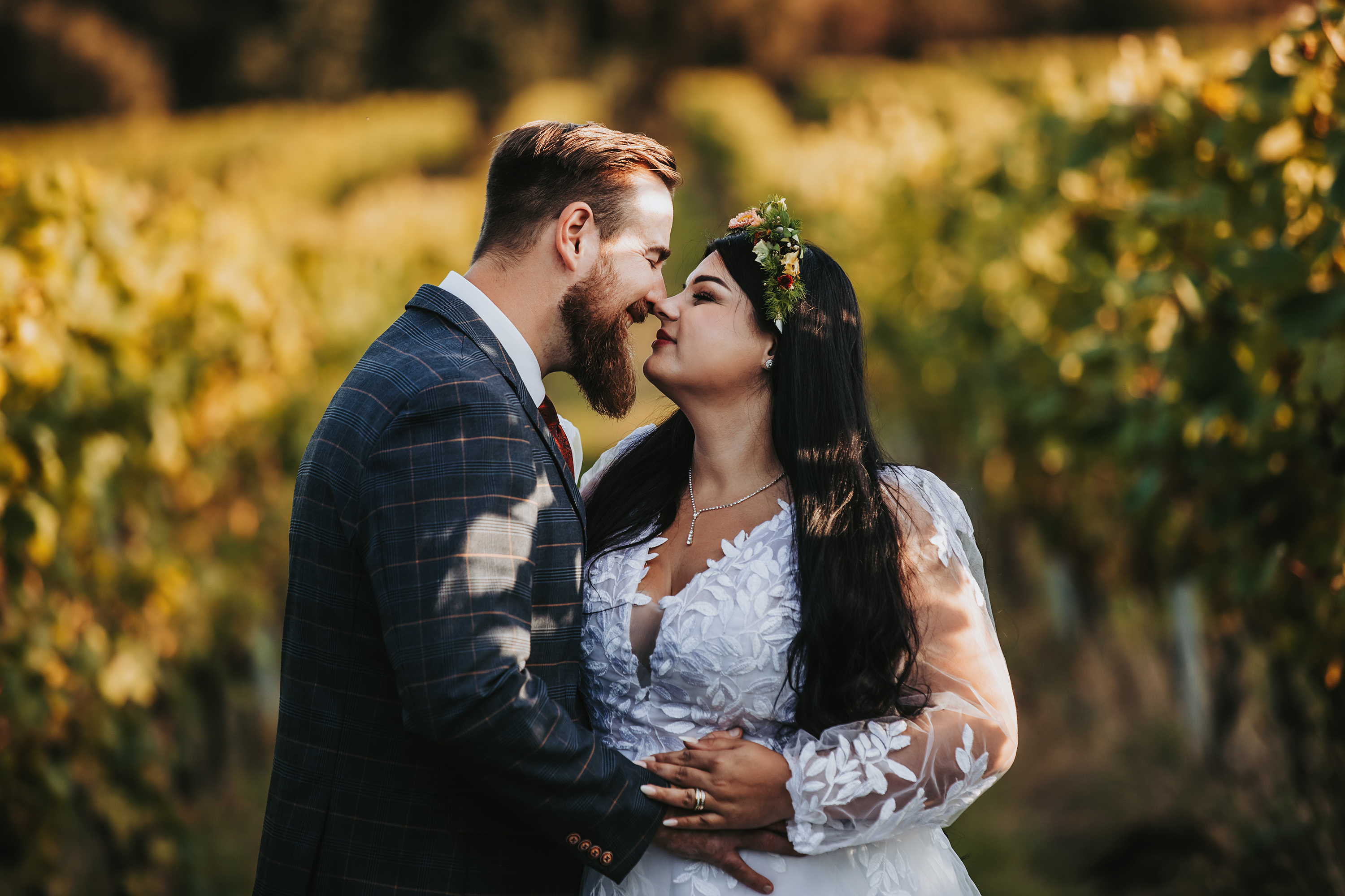 Romantic outdoor wedding portrait: bride in white floral lace and groom in navy plaid suit embrace in golden daylight among blurred vineyard greenery.