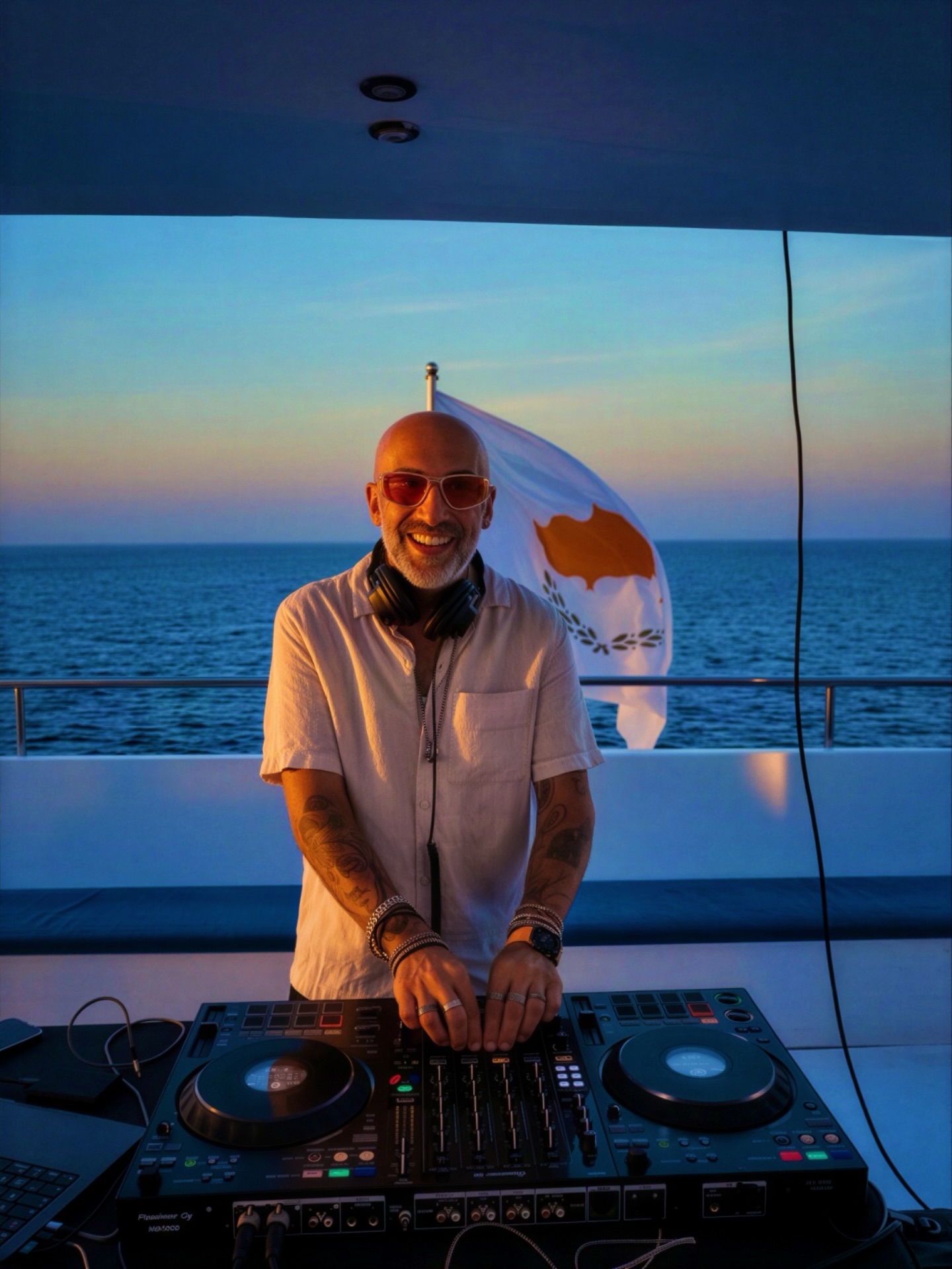 Smiling DJ on a yacht, mixing on twin Pioneer decks at sunset, with ocean backdrop, Cyprus flag, and sleek modern service vibe.