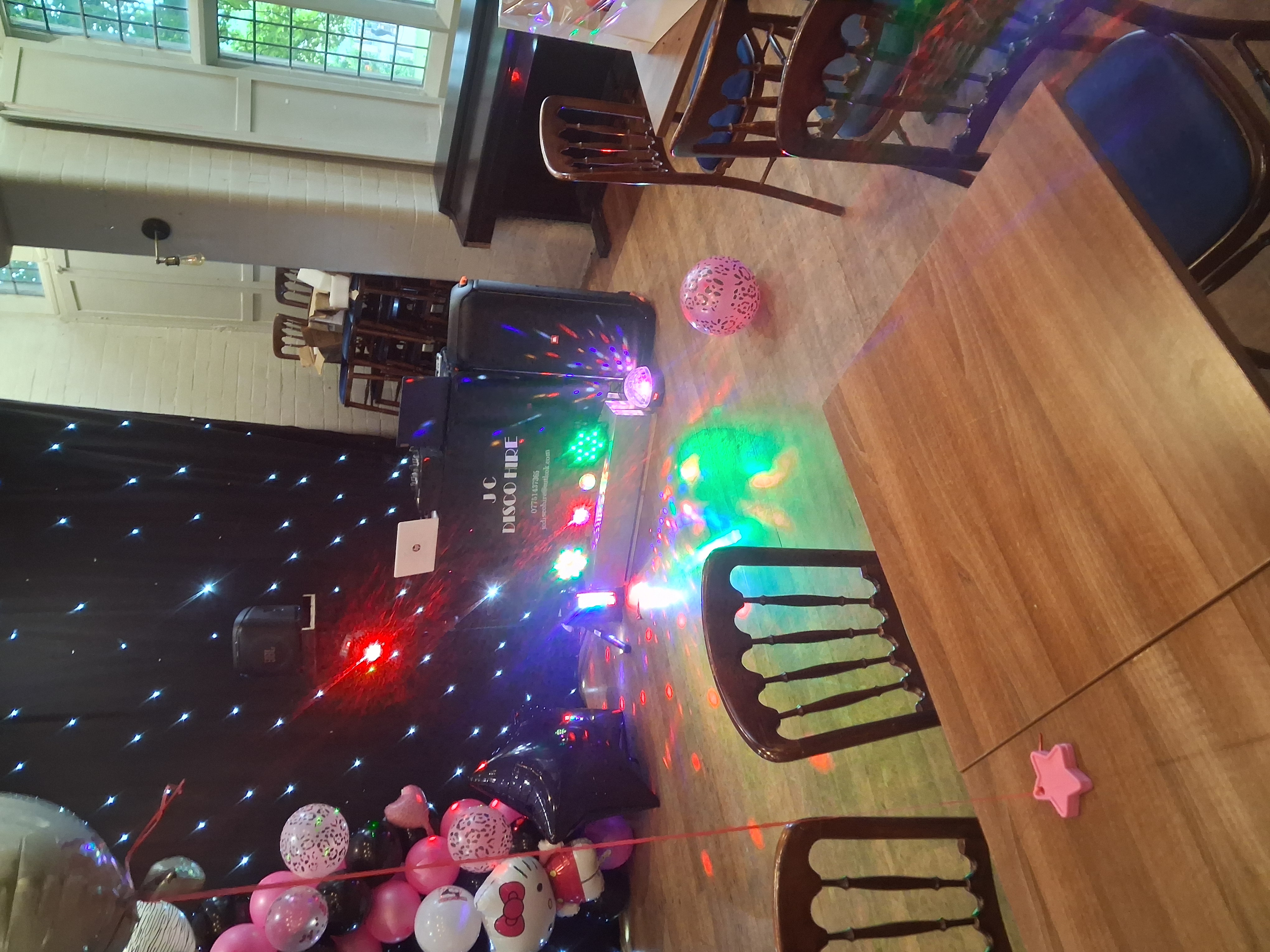 Indoor party venue with DJ booth, black star backdrop, pink balloons, and colorful lights beside rustic wooden tables and chairs.