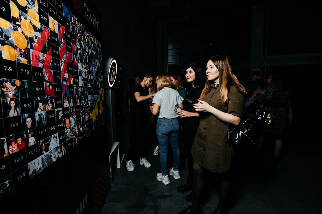 A vibrant event scene features guests mingling near a photo collage wall in dim lighting, capturing lively moments and fun energy.