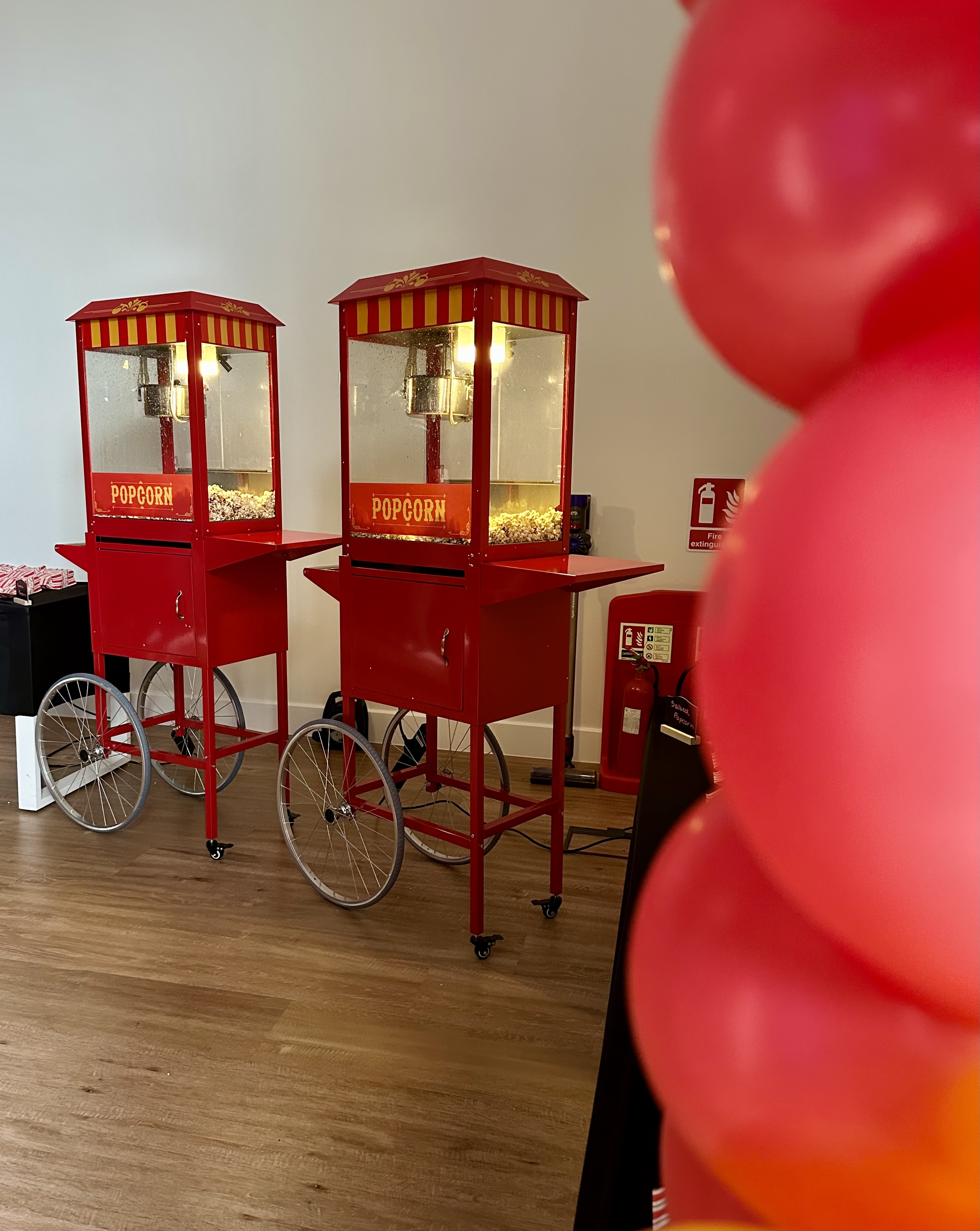 Two vintage red popcorn carts, illuminated by bright lights, stand on polished wooden flooring, evoking a festive, nostalgic atmosphere.