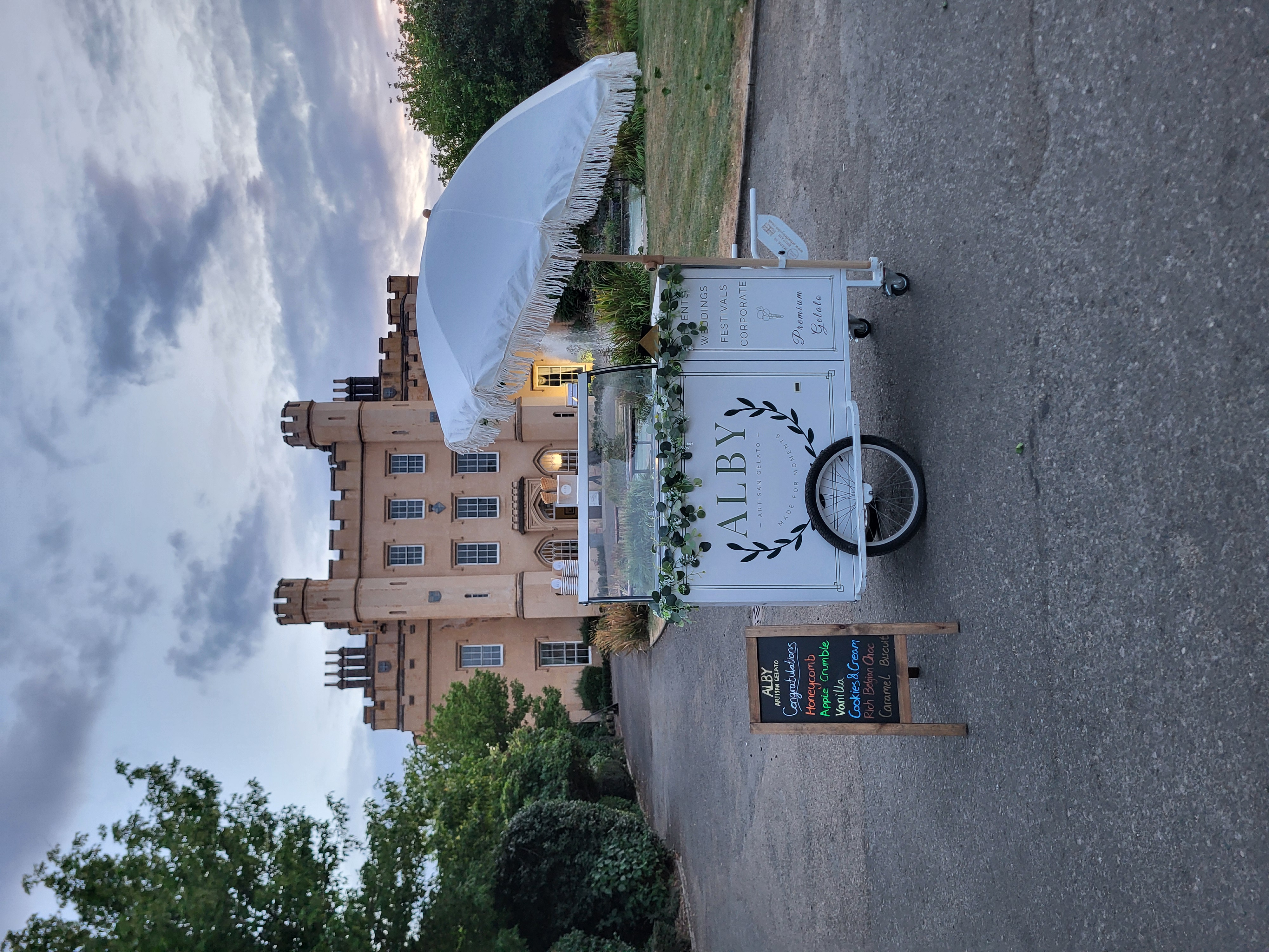 Elegant wedding gelato cart with white fringed parasol, chalkboard flavors, and castle backdrop, styled for outdoor catering in dusk light.