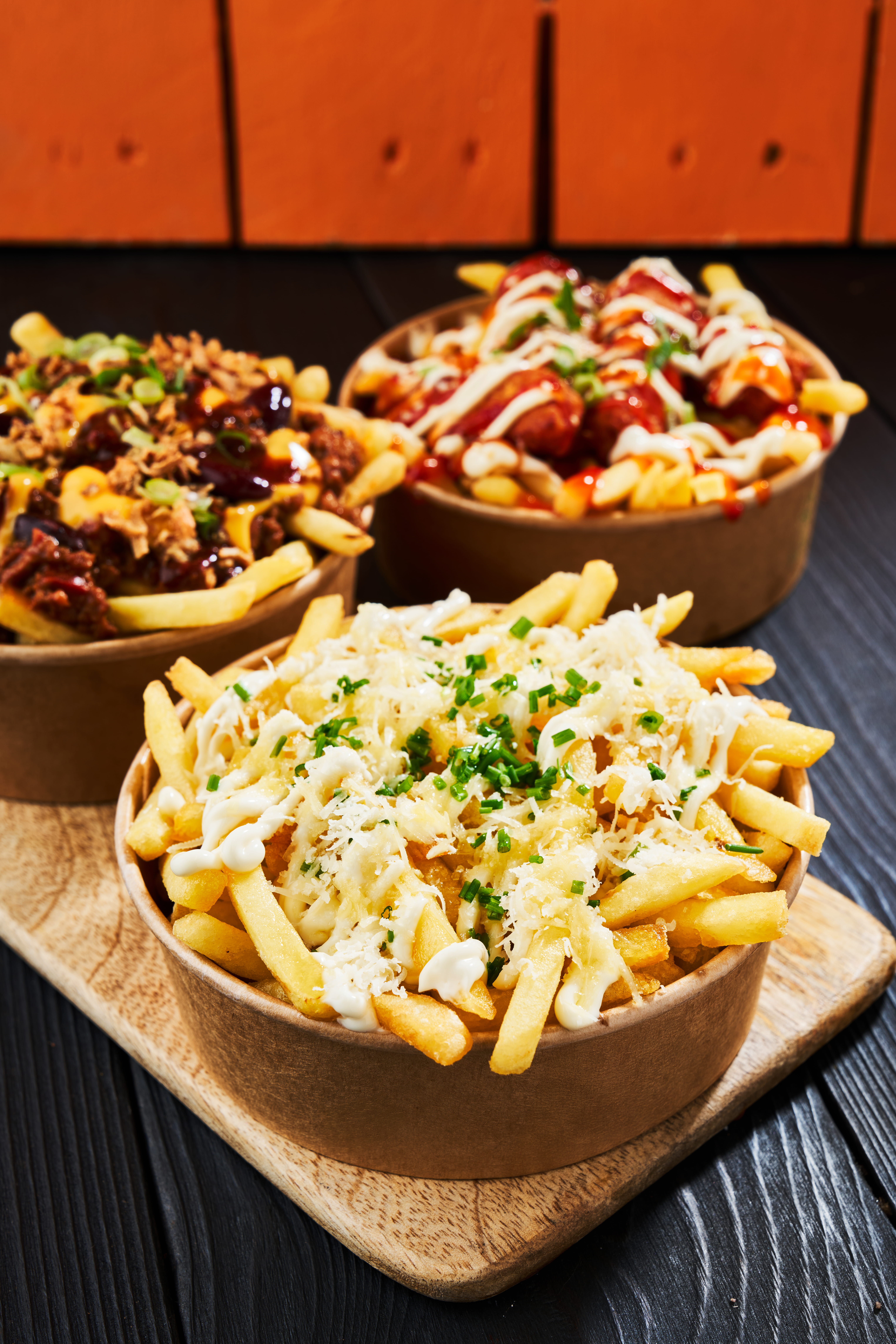 Three generous bowls of golden fries topped with various ingredients, including cheese, scallions, and sauces, on a wooden serving board.